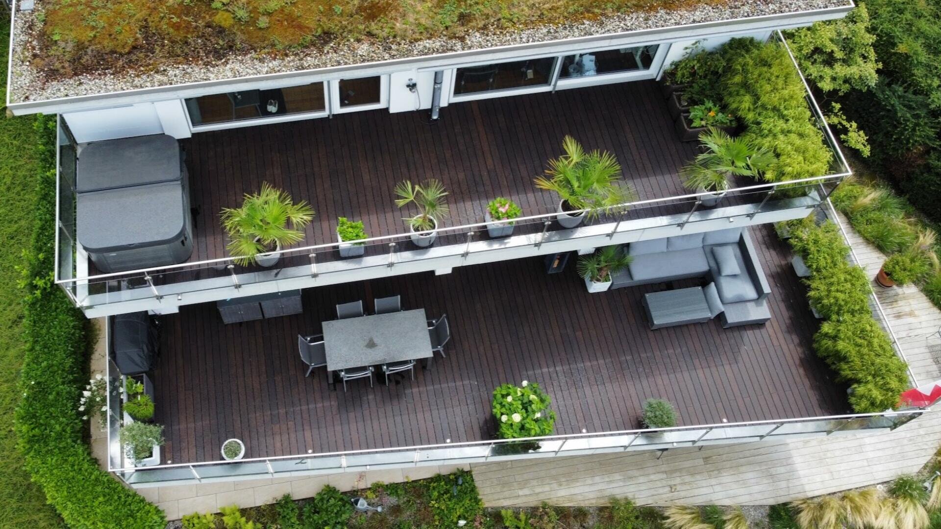 Luftaufnahme eines modernen Hausbalkons mit dunklem Holzfußboden, Gartenmöbeln, Topfpflanzen und Grünpflanzen. Der Balkon hat zwei Ebenen mit Ess- und Sitzbereichen, umgeben von einem Garten und einem Holzsteg.