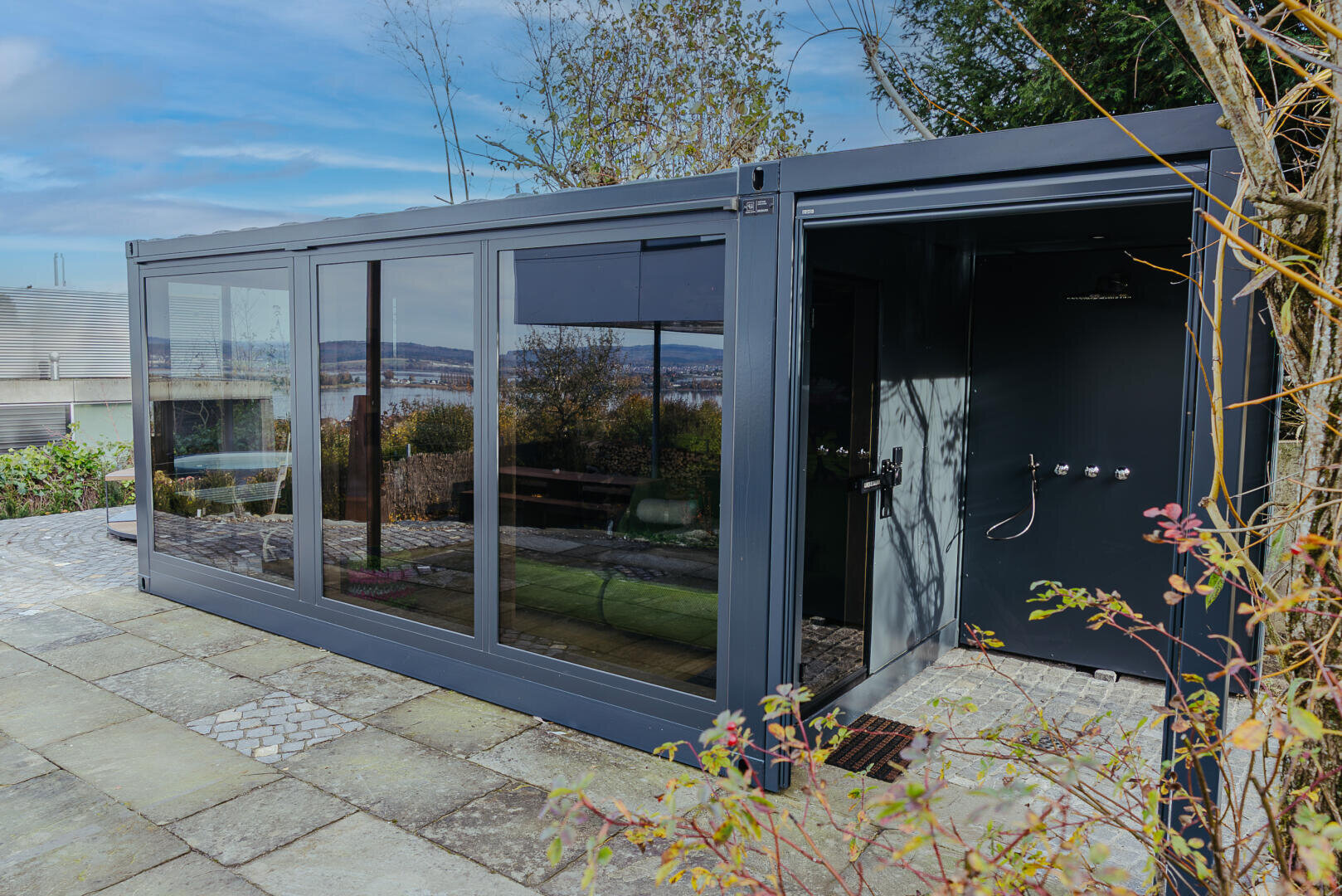 A modern garden room with large glass doors and windows stands on a stone terrace surrounded by plants. An outdoor shower can be seen on one side, and in the background are trees under a partly cloudy sky.