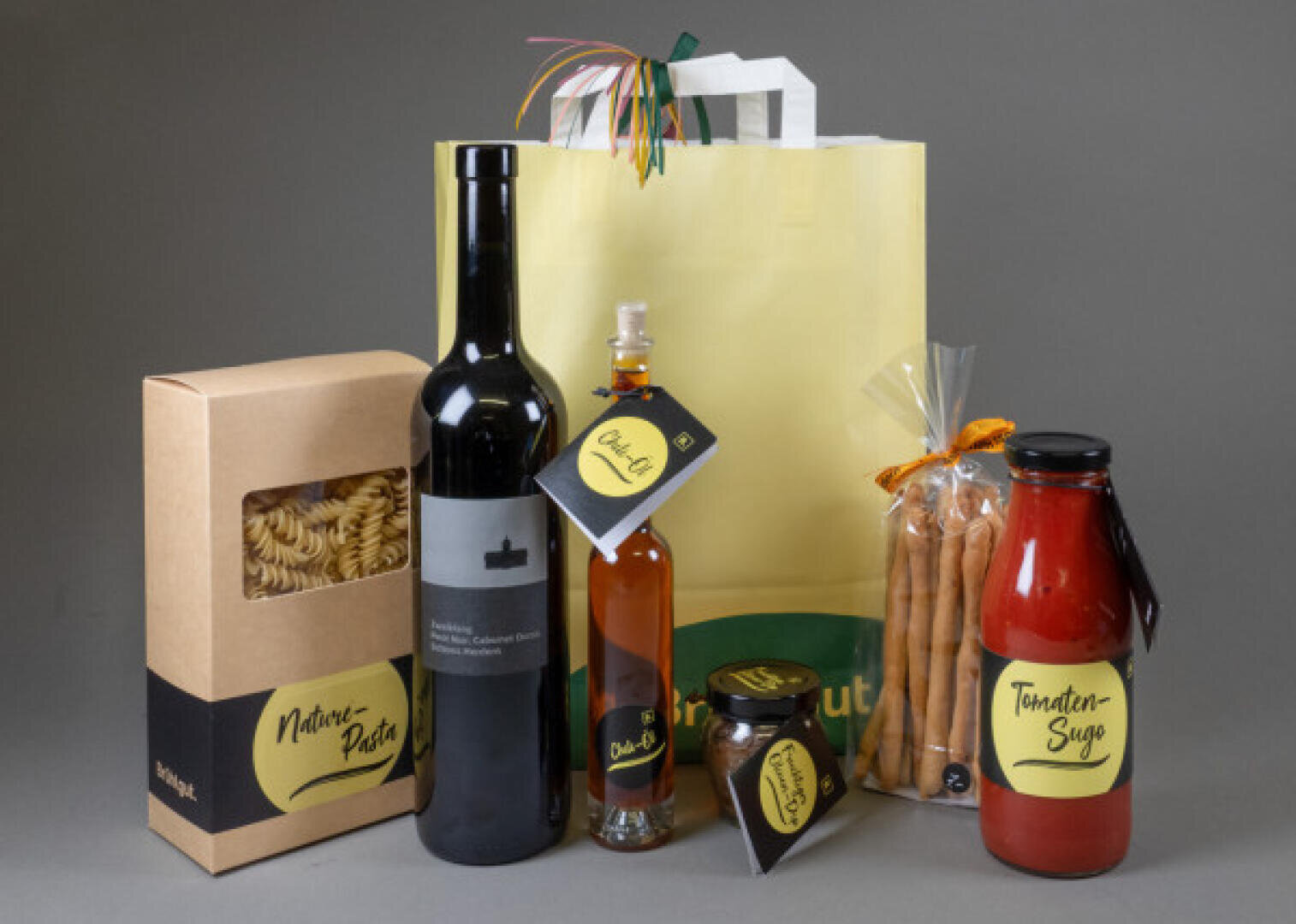 A selection of gourmet items, including a bottle of wine, a pasta box, a bottle of olive oil, breadsticks, a jar of tomato sauce and a small jar, all with matching yellow and black labels, arranged next to a yellow gift bag.