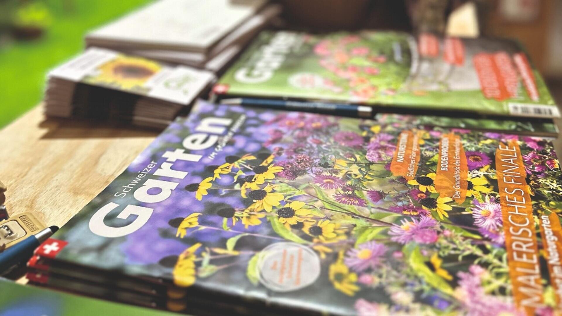 A close-up of gardening magazines and brochures on a wooden table, with colorful covers featuring flowers and the title Garten visible. Pens and blurred greenery are in the background.