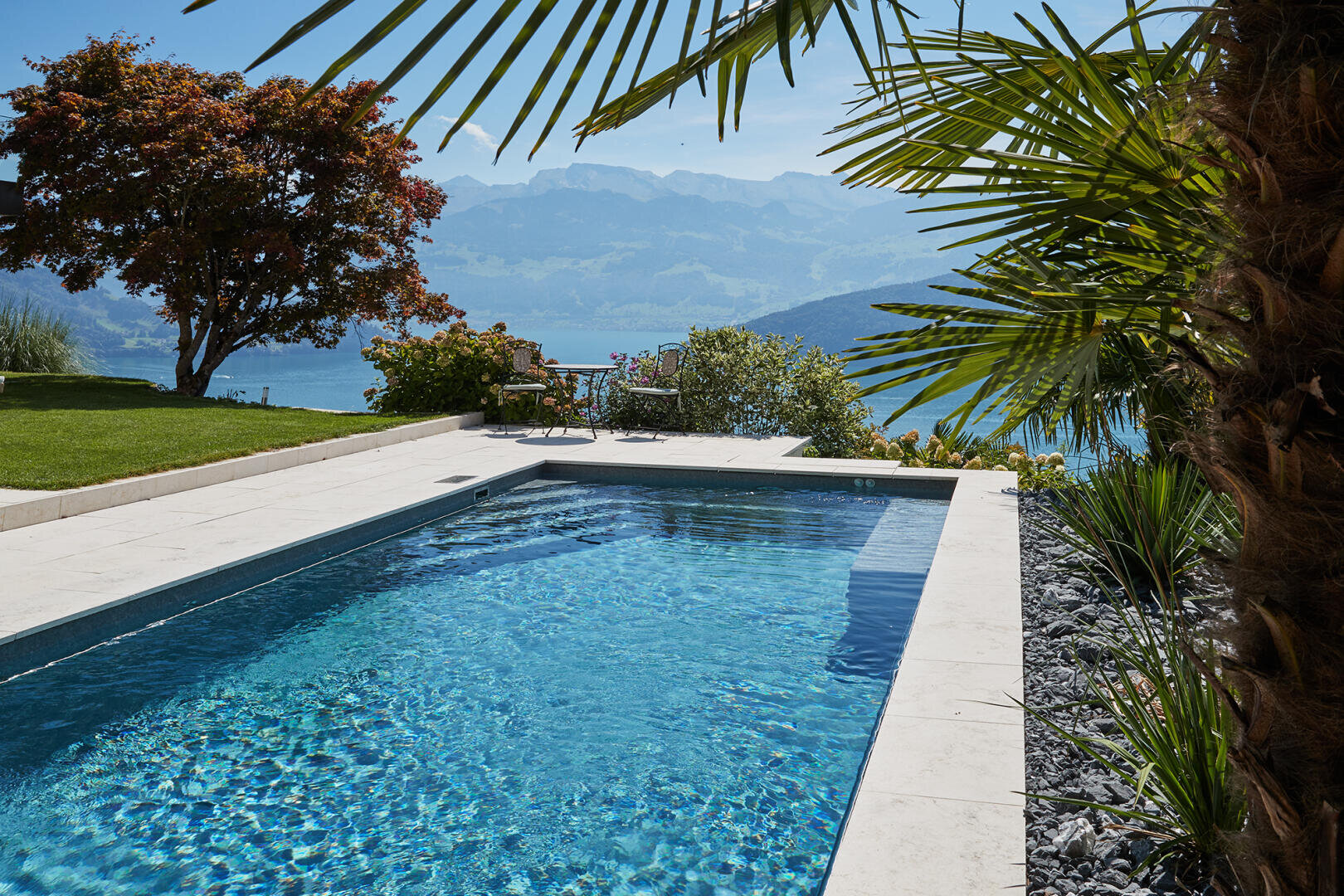 Ein klarer blauer Swimmingpool ist von Steinfliesen und üppigem Grün umgeben, darunter Palmen und blühende Sträucher, mit Blick auf einen See und Berge im Hintergrund unter einem sonnigen Himmel.
