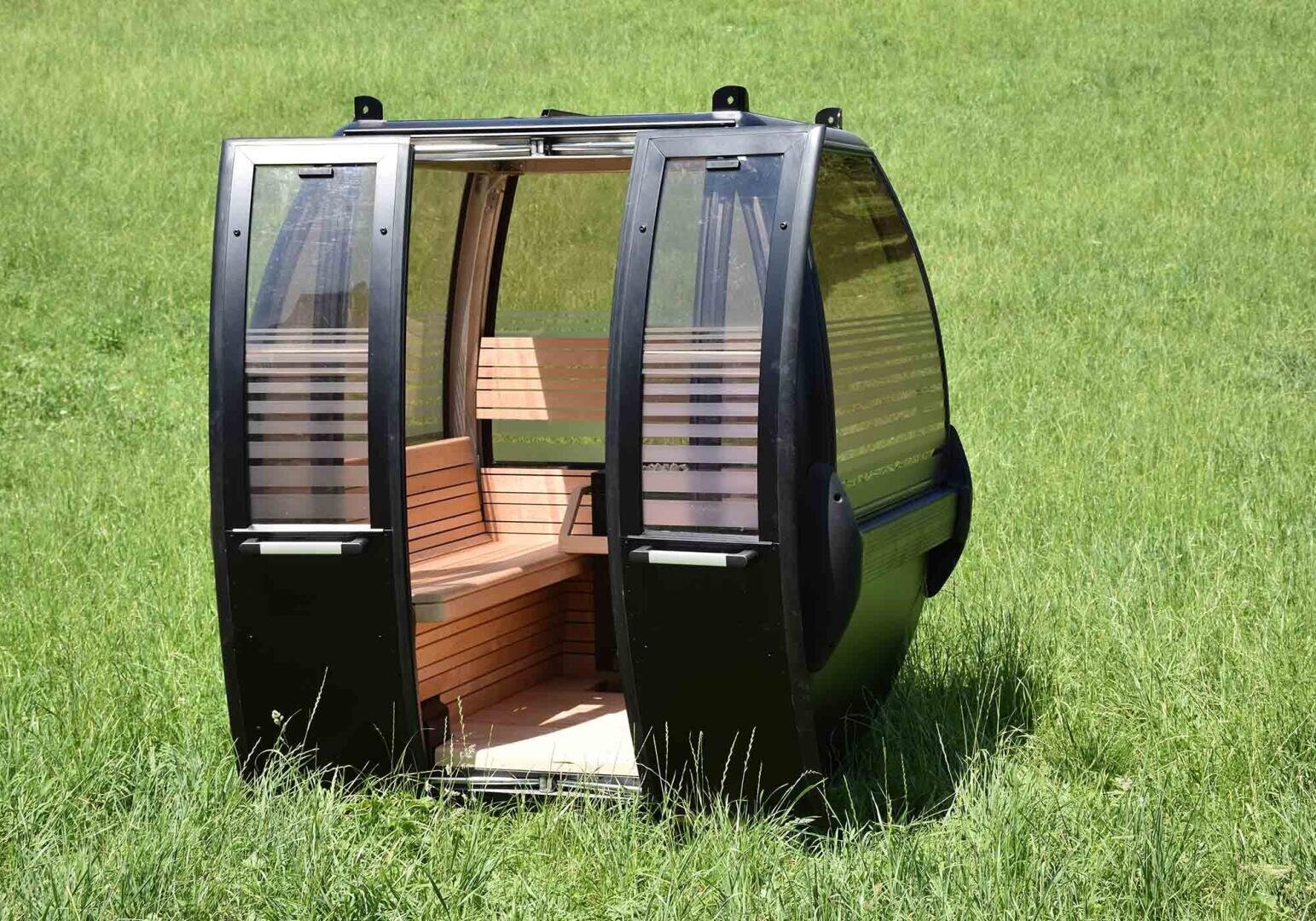 Eine schwarze, gläserne Gondelkabine mit hölzernen Sitzgelegenheiten steht auf grünem Gras in einem offenen Feld, losgelöst von jeglicher Kabel- oder Stützstruktur.