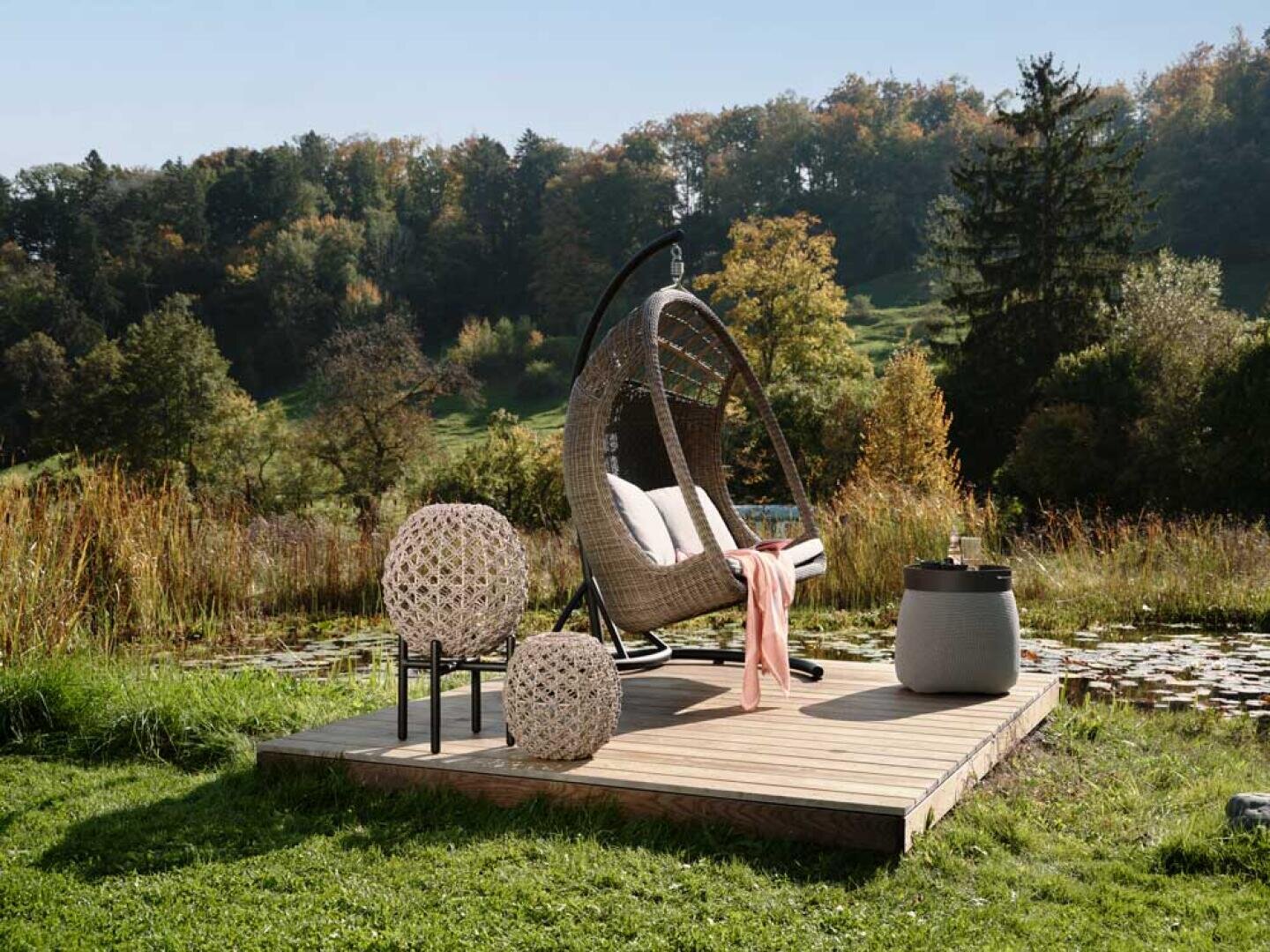 Eine gemütliche Sitzecke im Freien mit einem hängenden Korbstuhl, einem kleinen Tisch und zwei dekorativen Laternen auf einer Holzplattform an einem Teich, umgeben von üppigem Grün und Bäumen.