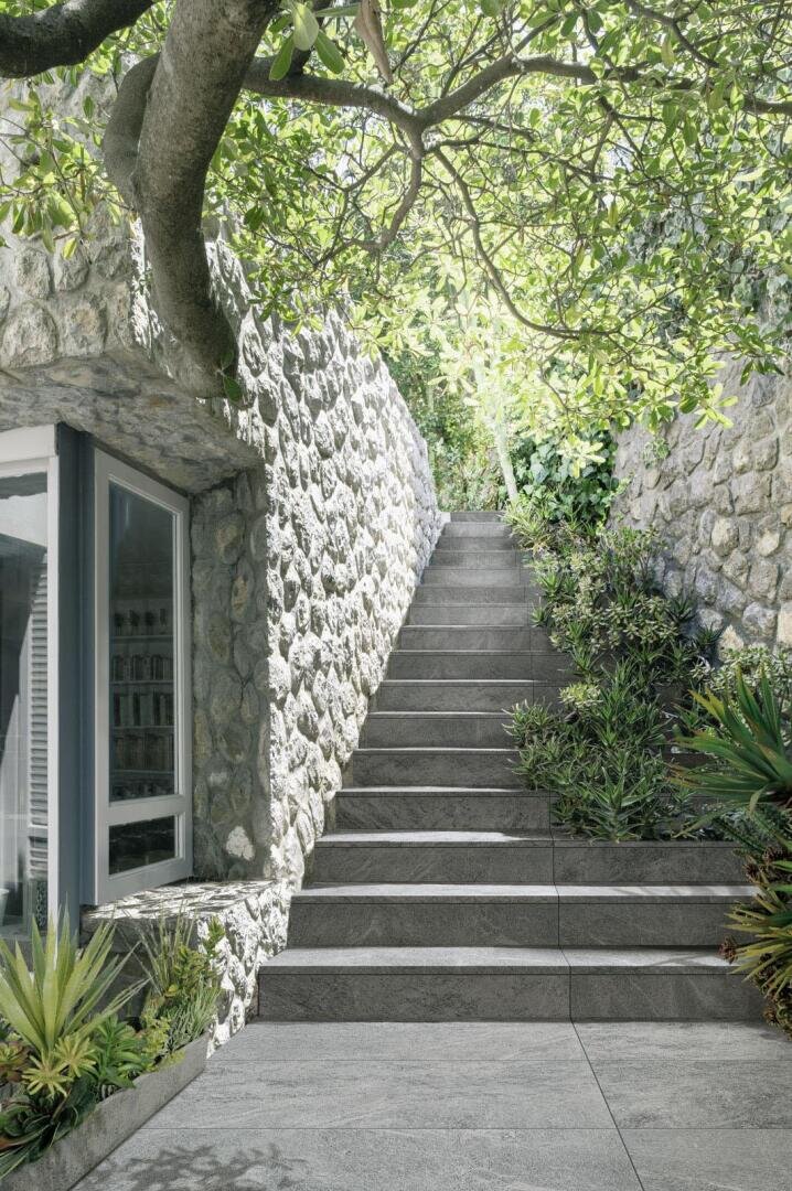 Eine Steintreppe, die von Steinmauern und üppigem Grün gesäumt ist, führt im Freien unter einem belaubten Baum neben einem Gebäude mit großen Fenstern hinauf. Das Sonnenlicht fällt durch das Laub und schafft eine ruhige und einladende Atmosphäre.