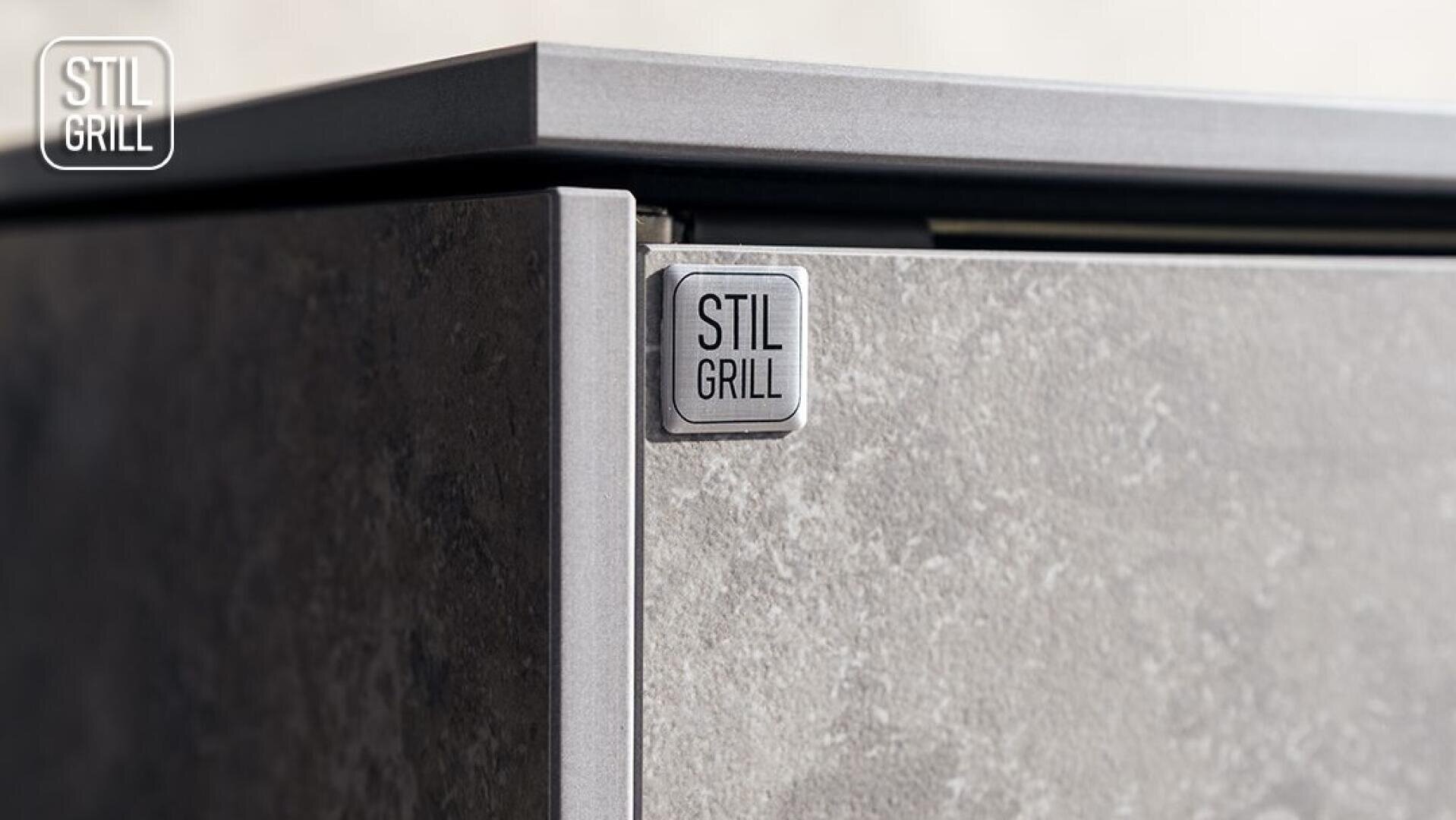 Close-up of the corner of a gray appliance with a stone structure and a metal plaque with the inscription STIL GRILL on the front. The STIL GRILL logo can also be seen in the top left corner of the picture.