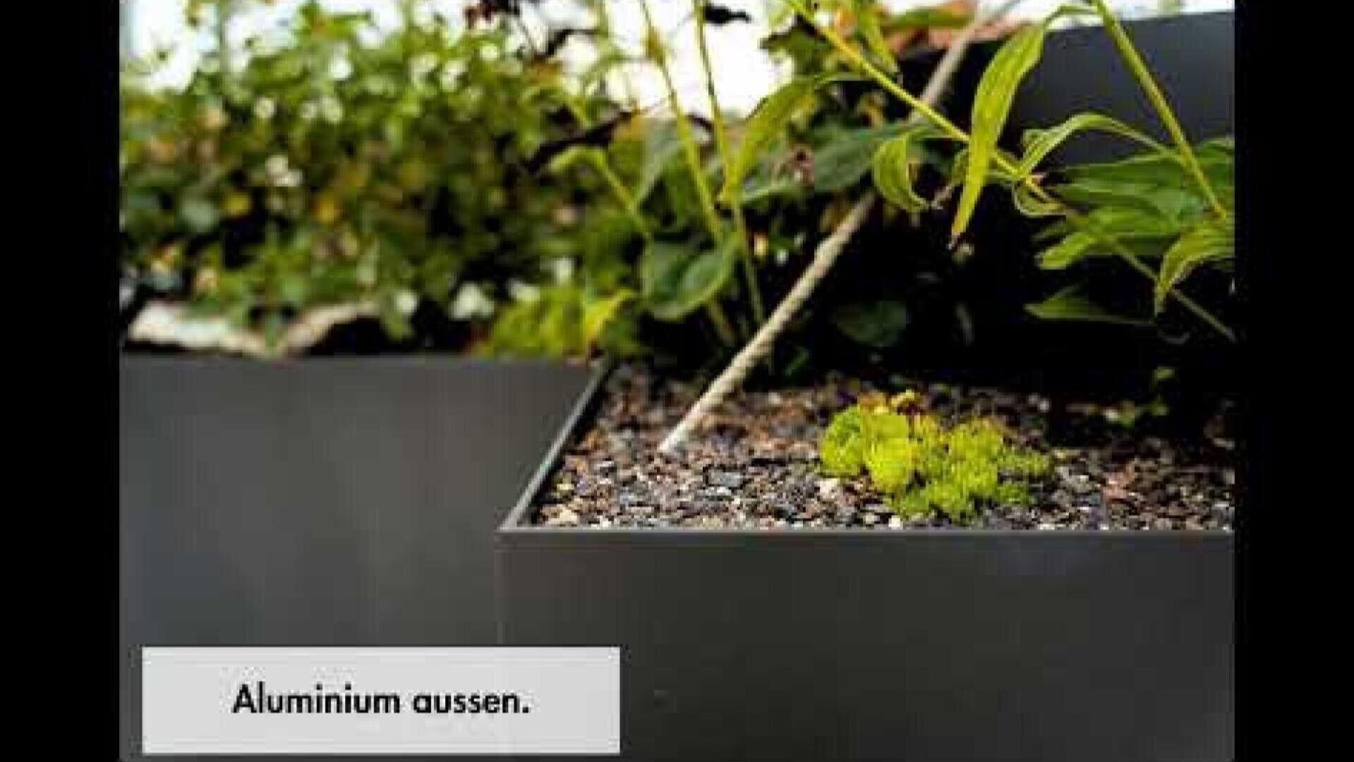 Nahaufnahme eines modernen rechteckigen Pflanzgefäßes aus dunklem Aluminium, gefüllt mit Erde und kleinen Grünpflanzen. Der deutsche Text unten links lautet "Aluminium aussen.".