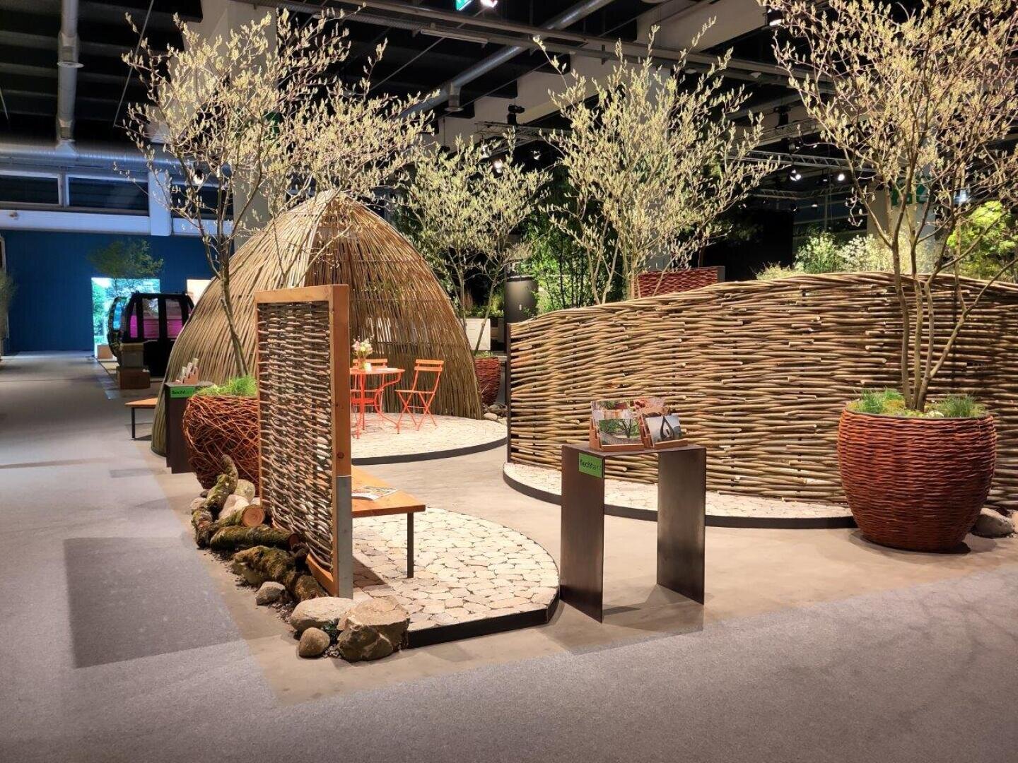 Eine Innenausstellung mit geflochtenen Weidenwänden, gewölbten Strukturen, Bäumen in großen Pflanzgefäßen, kleinen Sitzbereichen mit orangefarbenen Stühlen und Tischen sowie Steinwegen schafft eine natürliche und einladende Atmosphäre.