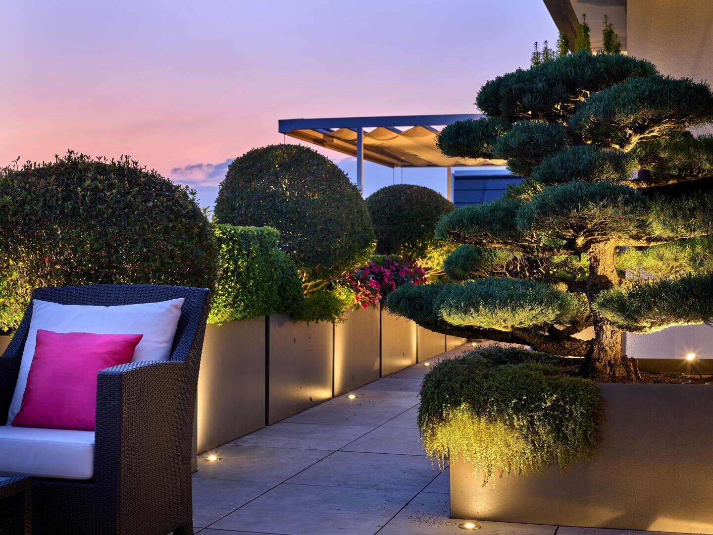 Eine moderne Dachterrasse bei Sonnenuntergang mit gepflegtem Grün in großen Pflanzgefäßen, sanfter Bodenbeleuchtung, einem Korbsessel mit rosa Kissen und einem gepflegten Baum schafft eine ruhige, einladende Atmosphäre.