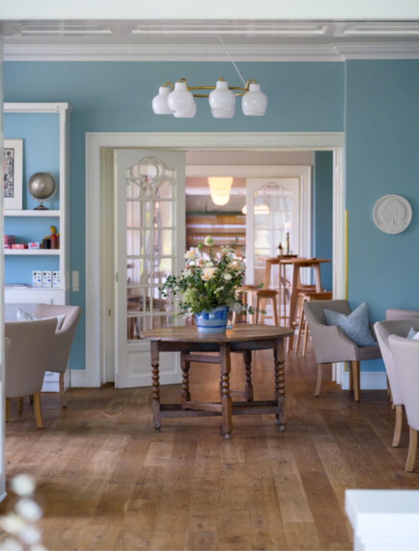 Ein gemütliches, elegantes Wohnzimmer mit blauen Wänden, Holzböden, hellbeigen Stühlen, einem runden Holztisch mit Blumenarrangement und doppelten Glastüren, die zu einem weiteren gut beleuchteten Raum mit weiteren Sitzgelegenheiten führen.