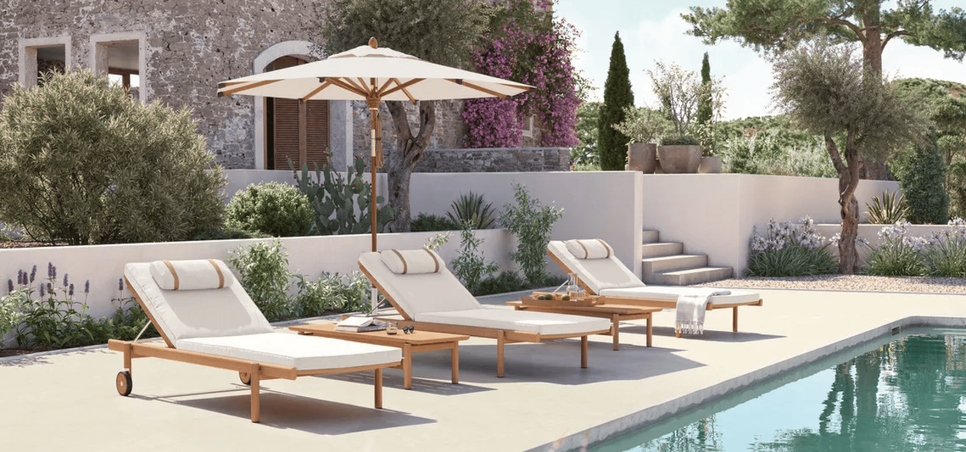 Three cushioned wooden lounge chairs with an umbrella are arranged beside a swimming pool. Behind them is a stone building with greenery, flowering vines, and a landscaped garden under a sunny sky.
