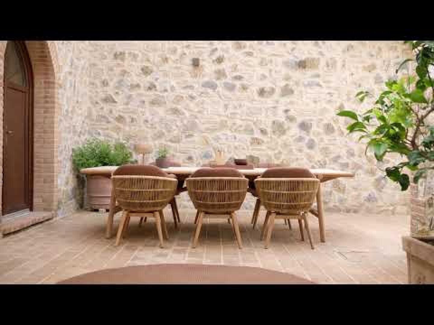 A rustic outdoor terrace with a stone wall, a wooden table and six wicker chairs. The area is surrounded by potted plants that create a cozy and inviting atmosphere.