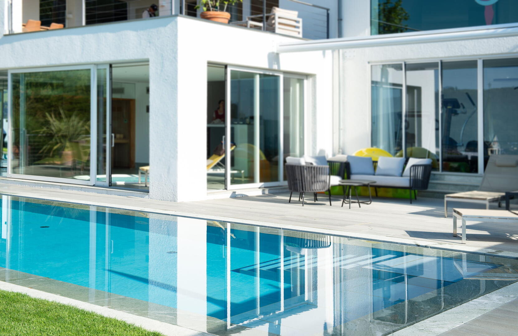 Modernes Haus mit großen Glasschiebetüren, die sich zu einer Terrasse mit Sitzgelegenheiten im Freien öffnen, neben einem klaren blauen Swimmingpool; üppiges grünes Gras umgibt den Bereich, und das Sonnenlicht erhellt die Szene.