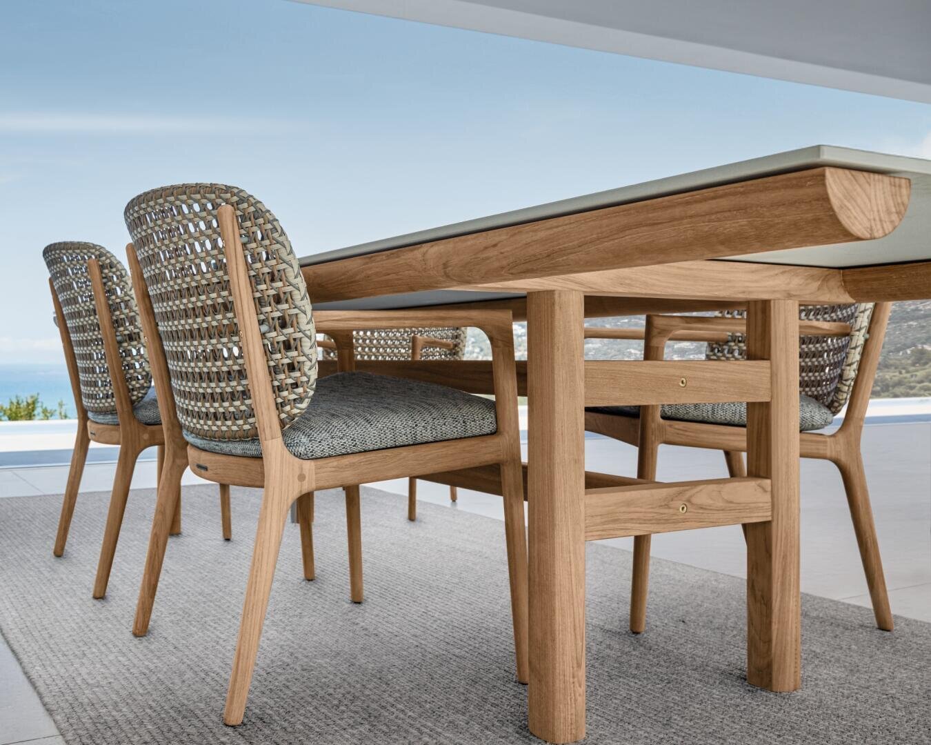 Nahaufnahme eines hölzernen Esstisches mit vier Stühlen mit geflochtener Rückenlehne auf einer Terrasse, mit Blick auf eine malerische Landschaft und den blauen Himmel im Hintergrund.