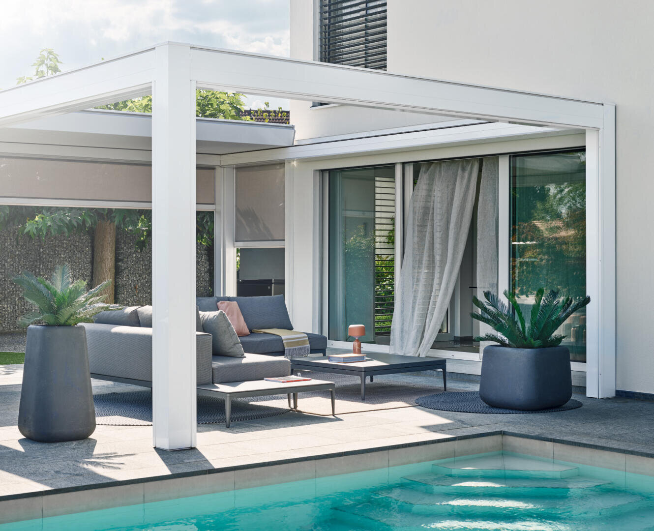 Moderne Außenterrasse mit grauen Möbeln und Topfpflanzen unter einer weißen Pergola neben einem Swimmingpool, mit teilweise geöffneten transparenten Vorhängen an den Glasschiebetüren, die ins Haus führen.