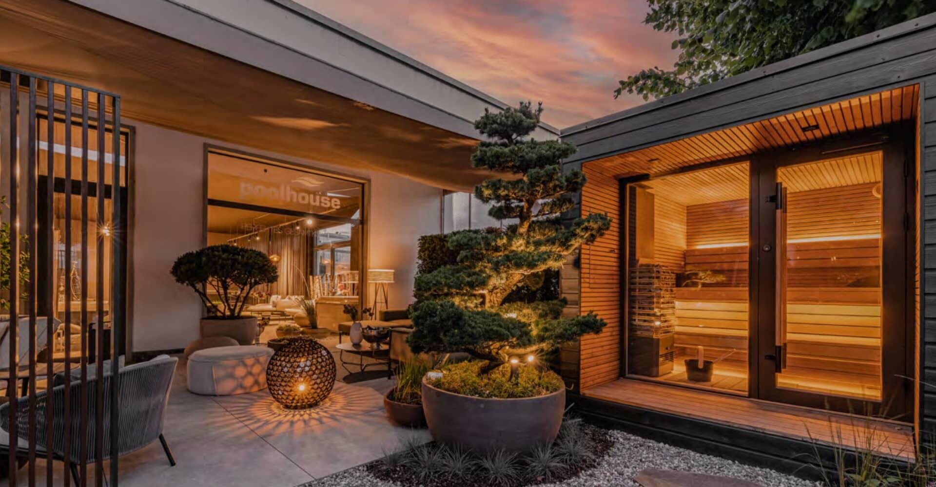 Eine moderne Außenterrasse bei Sonnenuntergang mit einer gemütlichen Sitzecke, Topfpflanzen, warmer Beleuchtung und einem hölzernen Saunaraum mit Glastüren.