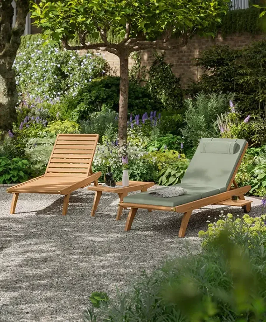 Two wooden lounge chairs, one with a green cushion and headrest, sit beside a small wooden side table with a vase and cup in a lush garden with trees, flowers, and leafy plants on a gravel area.