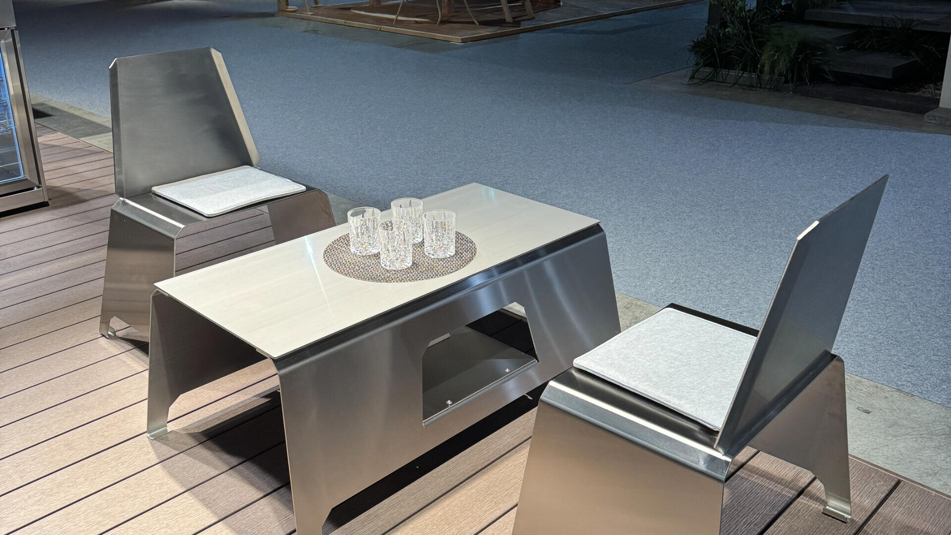 A modern metal table with four clear glasses on a round placemat is flanked by two matching metal chairs with light gray seat cushions, set on a wooden deck in an indoor space.