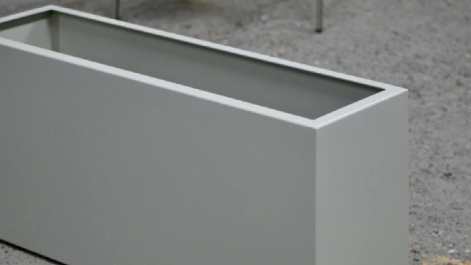 A rectangular, light gray planter box sits on a rough, gray concrete surface. The planter is empty and modern in design, with clean, straight lines.