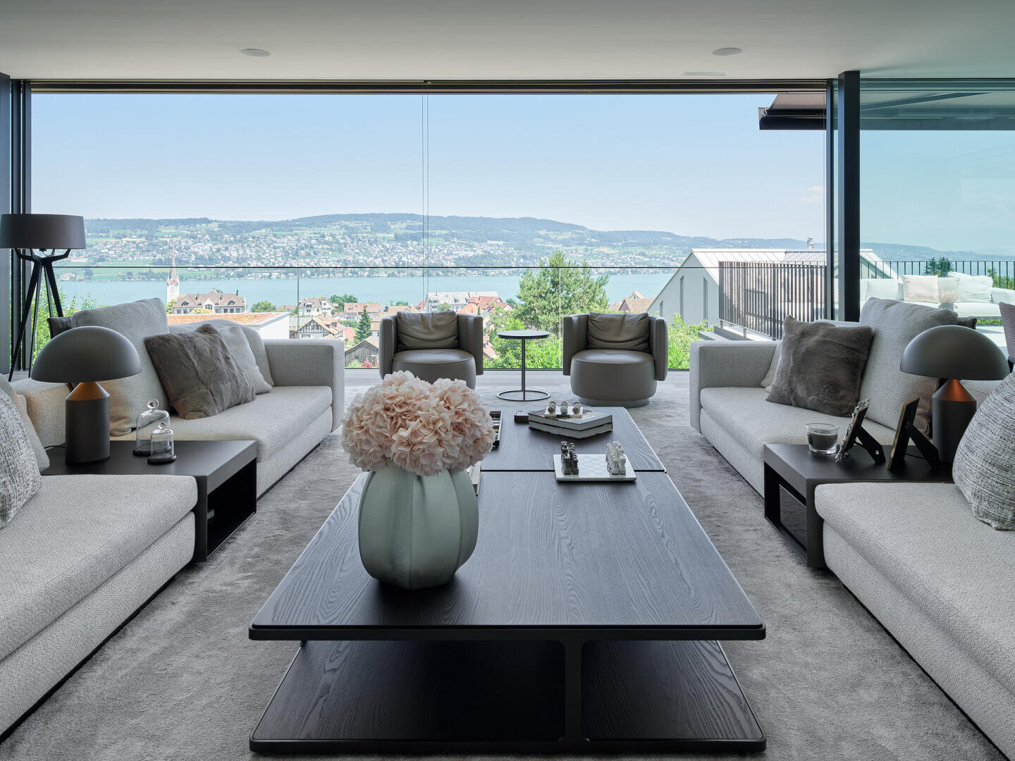 Modern living room with two beige sofas, a black coffee table, decorative items and large windows offering a panoramic view of a lake, the city and distant hills under a blue sky.