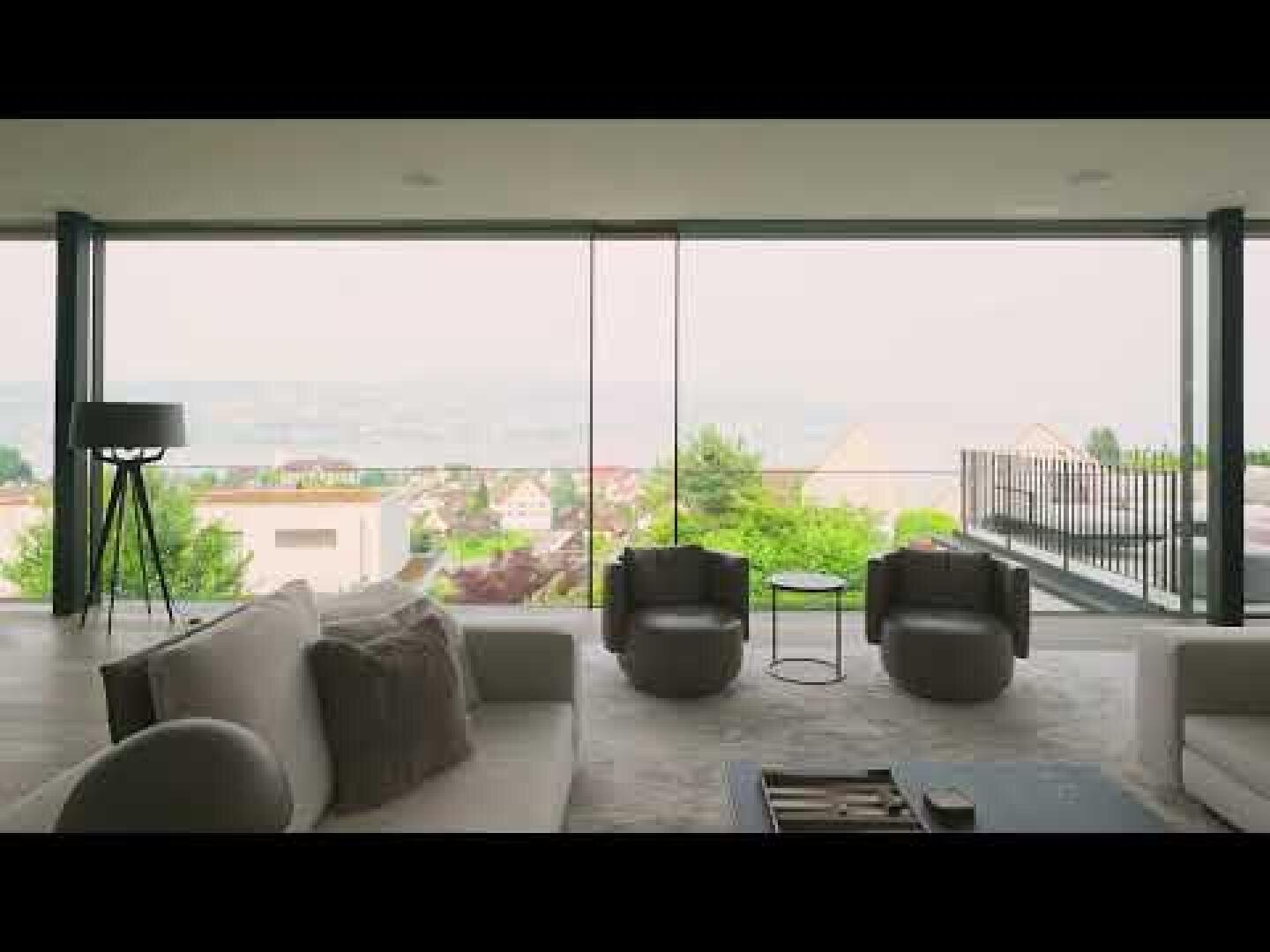 A modern living room with large floor-to-ceiling windows offering a picturesque, hazy view of rooftops, greenery and distant hills. The room has sofas in neutral tones, armchairs, a floor lamp and a balcony.