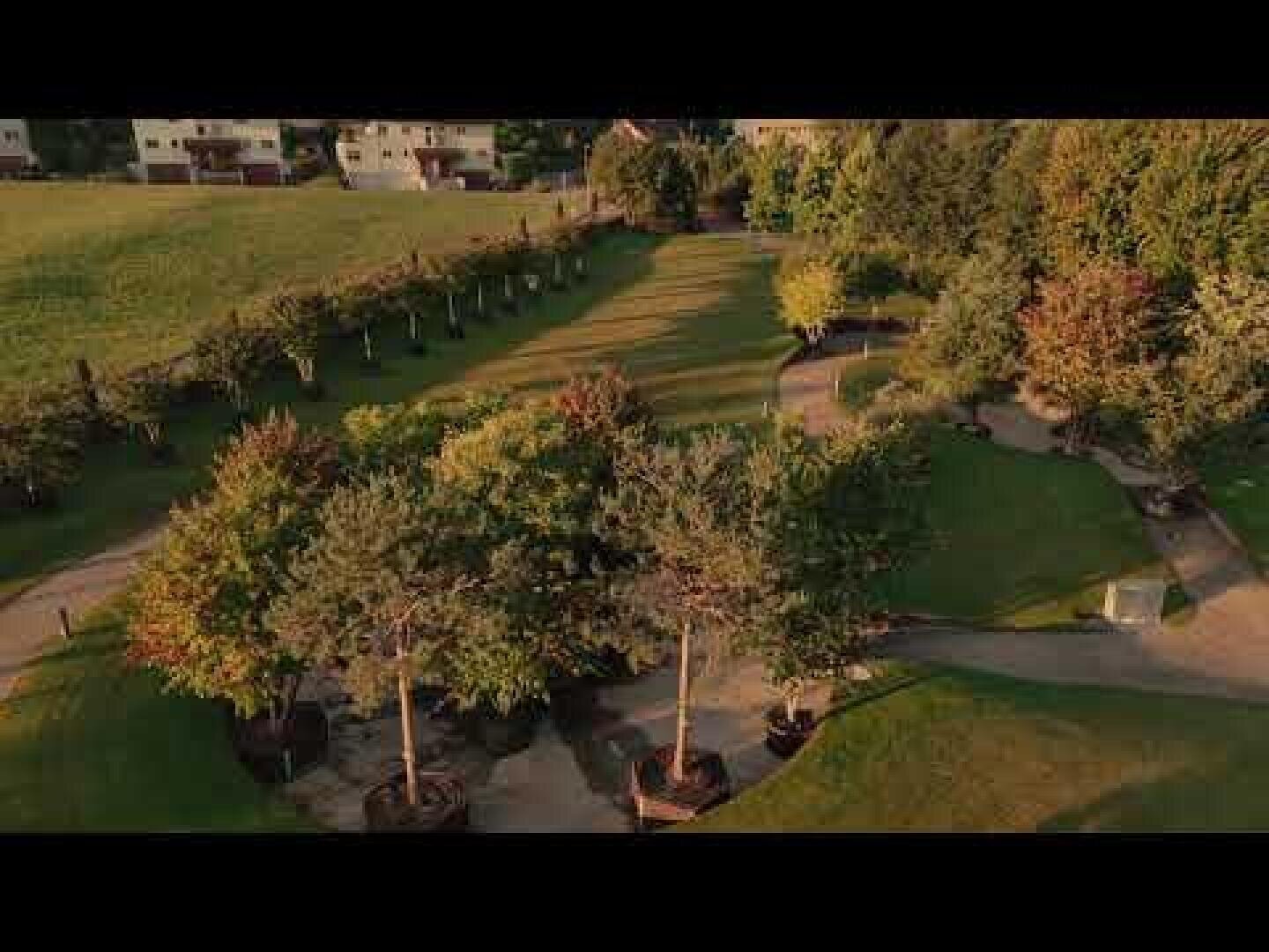 Luftaufnahme eines Landschaftsparks mit verschlungenen Wegen, grünen Rasenflächen und Baumgruppen, umgeben von einem Feld und Wohnhäusern im Hintergrund.