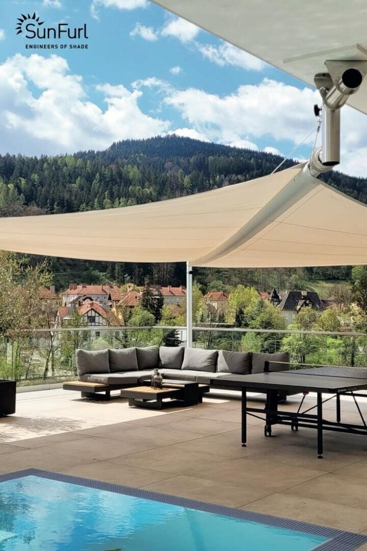 Eine moderne Außenterrasse mit beigen Sonnensegeln, einem Sitzbereich, einem Tisch und einem Pool blickt auf eine malerische Landschaft mit Bäumen, Hügeln und Dächern unter einem teilweise bewölkten Himmel. Das SunFurl-Logo erscheint in der oberen linken Ecke.