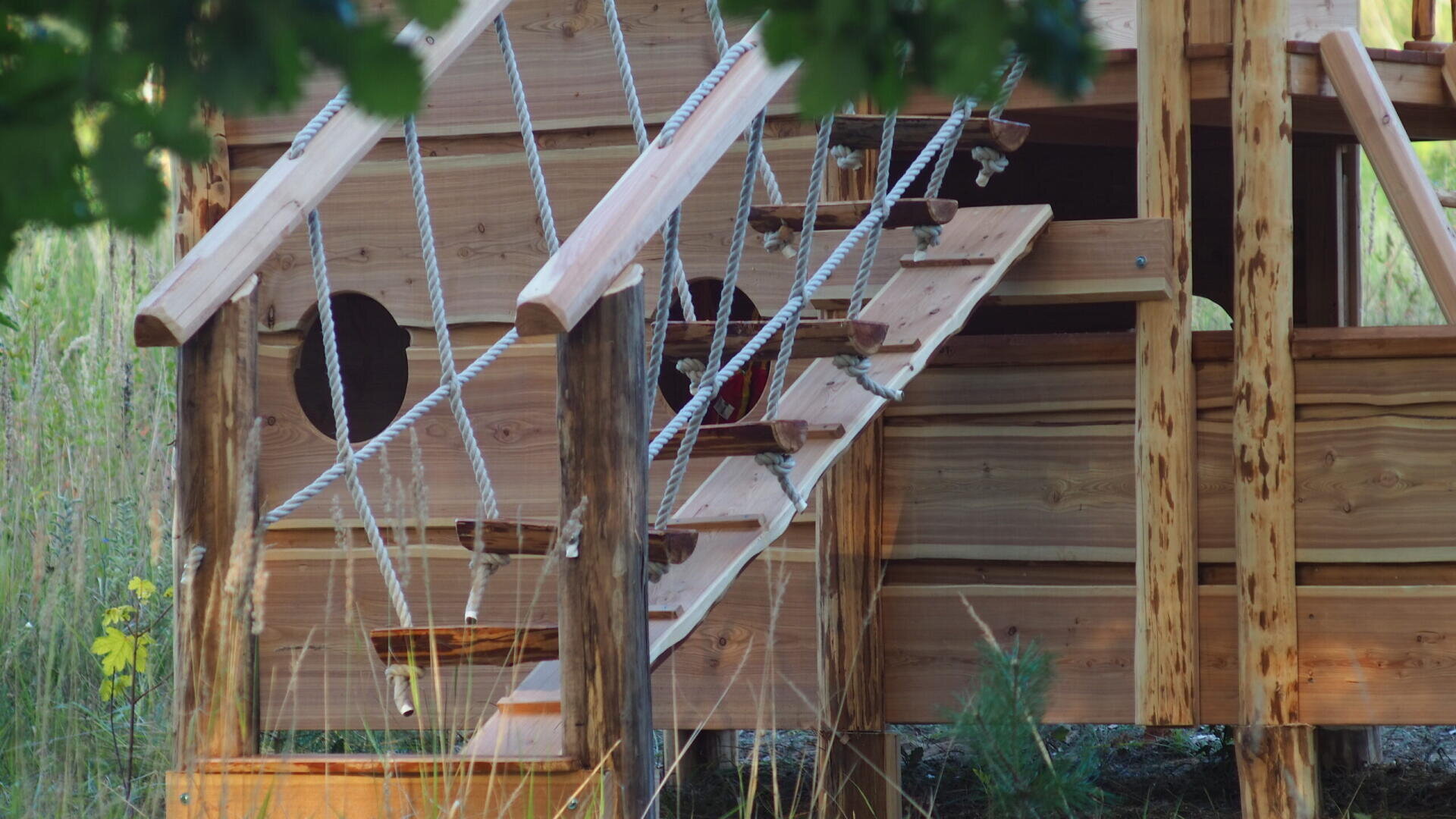 A close-up of a wooden play structure or treehouse with rope railings and a small wooden ladder, set outdoors among grass and trees.