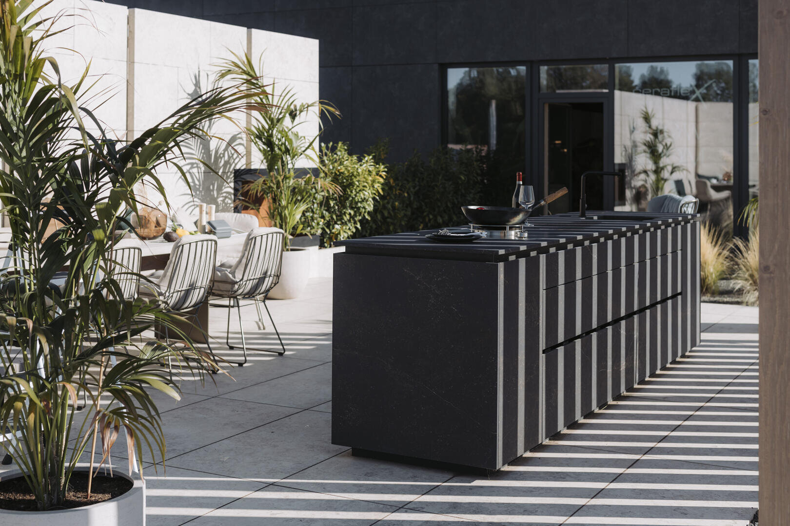 Moderne Außenterrasse mit einer eleganten schwarzen Kücheninsel, einem Esstisch mit Stühlen, Topfpflanzen und Schatten von Dachlatten, die Streifenmuster auf dem gefliesten Boden erzeugen.