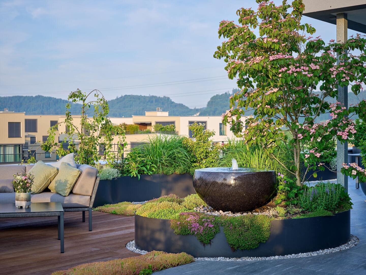 Ein moderner Dachgarten mit üppigem Grün, einem blühenden Baum, einem runden schwarzen Wasserbrunnen und einem gemütlichen Sitzbereich mit Kissen. Im Hintergrund sind moderne Gebäude und grüne Hügel zu sehen.