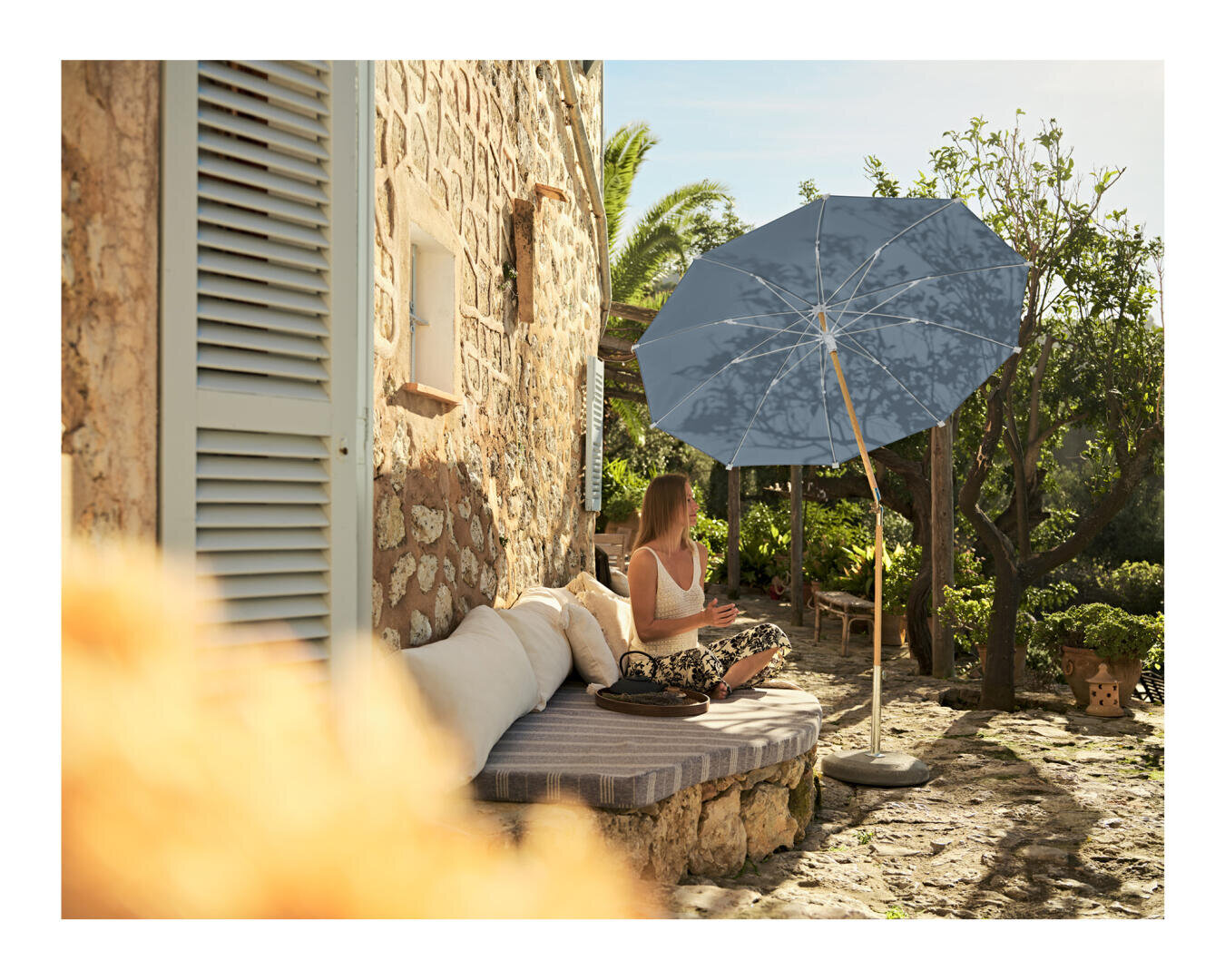 Eine Frau sitzt an einem sonnigen Tag auf einer gepolsterten Bank neben einem Steinhaus unter einem großen blauen Regenschirm, umgeben von Bäumen und Pflanzen.