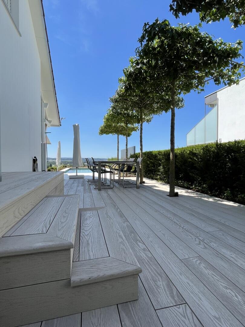 Eine moderne Außenterrasse mit hellen Holzböden, Stufen, einem Esstisch mit Stühlen, beschnittenen Bäumen und Sonnenschirmen am Swimmingpool. Der strahlend blaue Himmel und der Blick in die Ferne vervollständigen die heitere Szenerie.