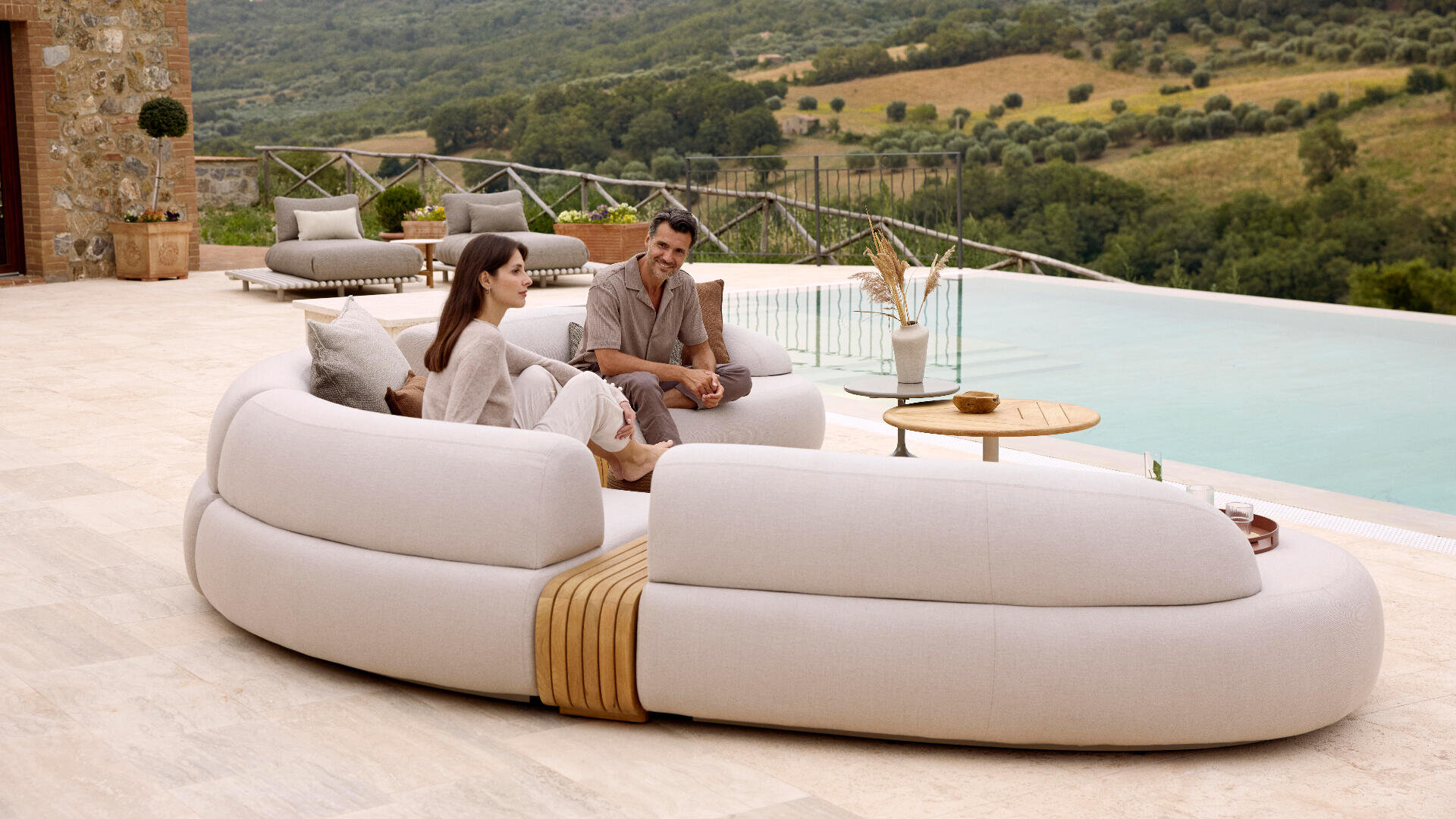 A man and woman sit and chat on modern white outdoor sofas beside a pool, with scenic green hills and trees in the background. A round wooden table with decor and a lounger are nearby.