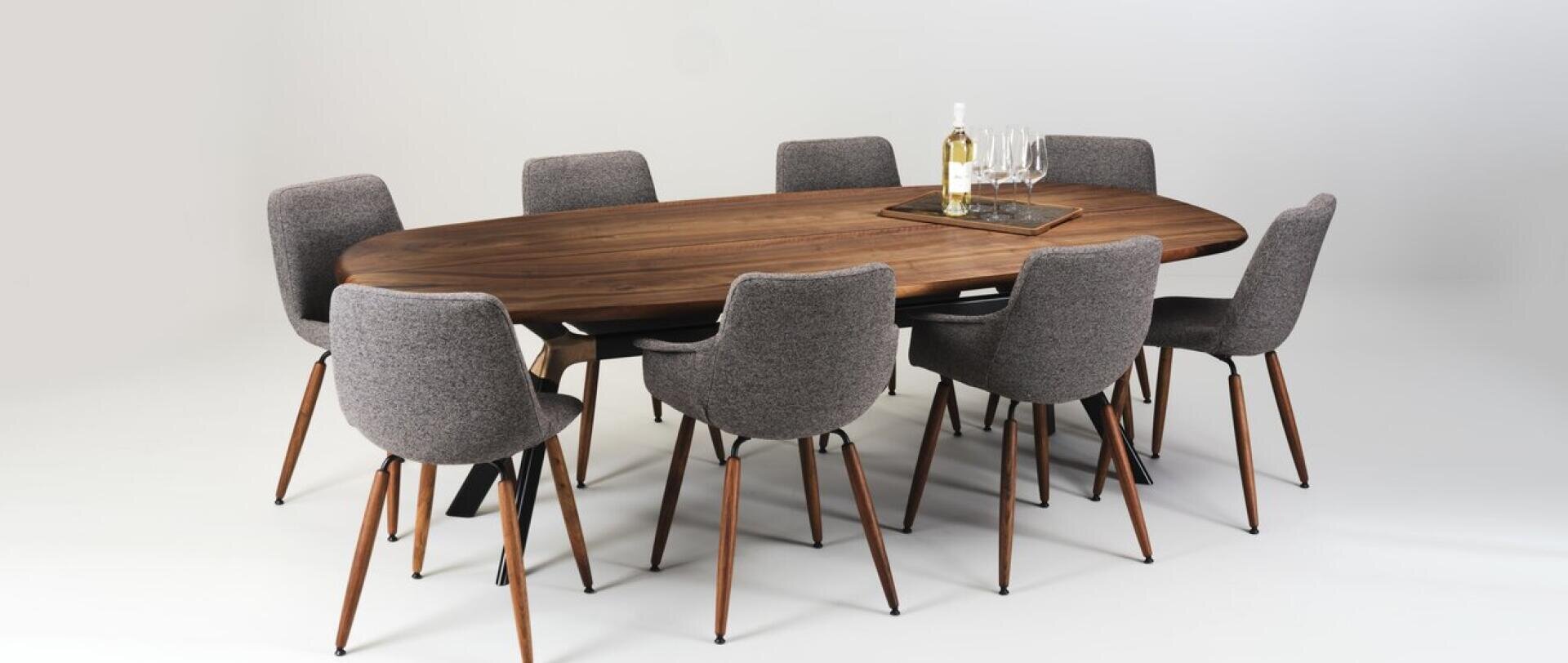 A modern oval wooden dining table with eight gray upholstered chairs. On the table is a tray holding a bottle of white wine and four wine glasses. The background is plain white.