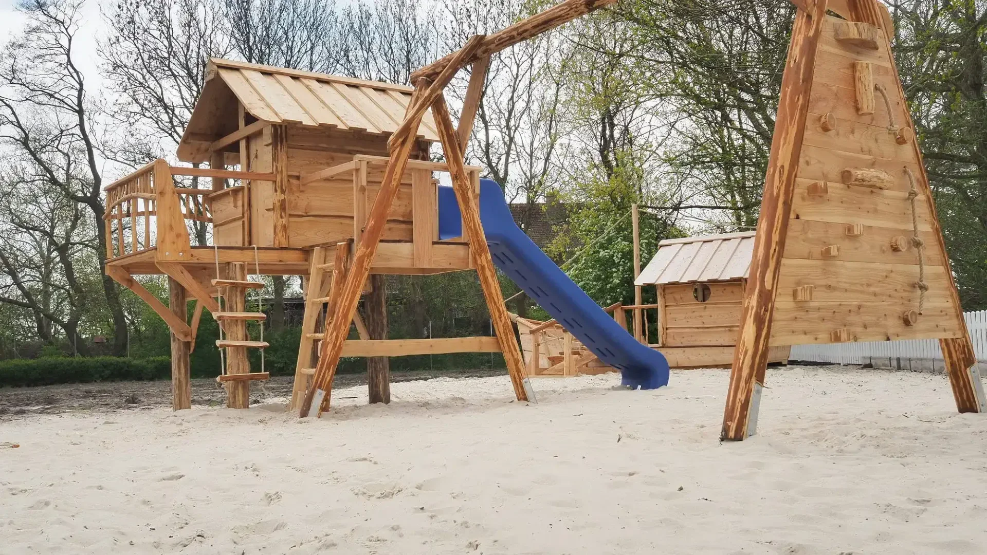 Ein hölzernes Spielplatzgerät mit einem Baumhaus, einer blauen Rutsche, Schaukeln und einer Strickleiter steht auf einer sandigen Fläche, umgeben von blattlosen Bäumen und Grün im Hintergrund.