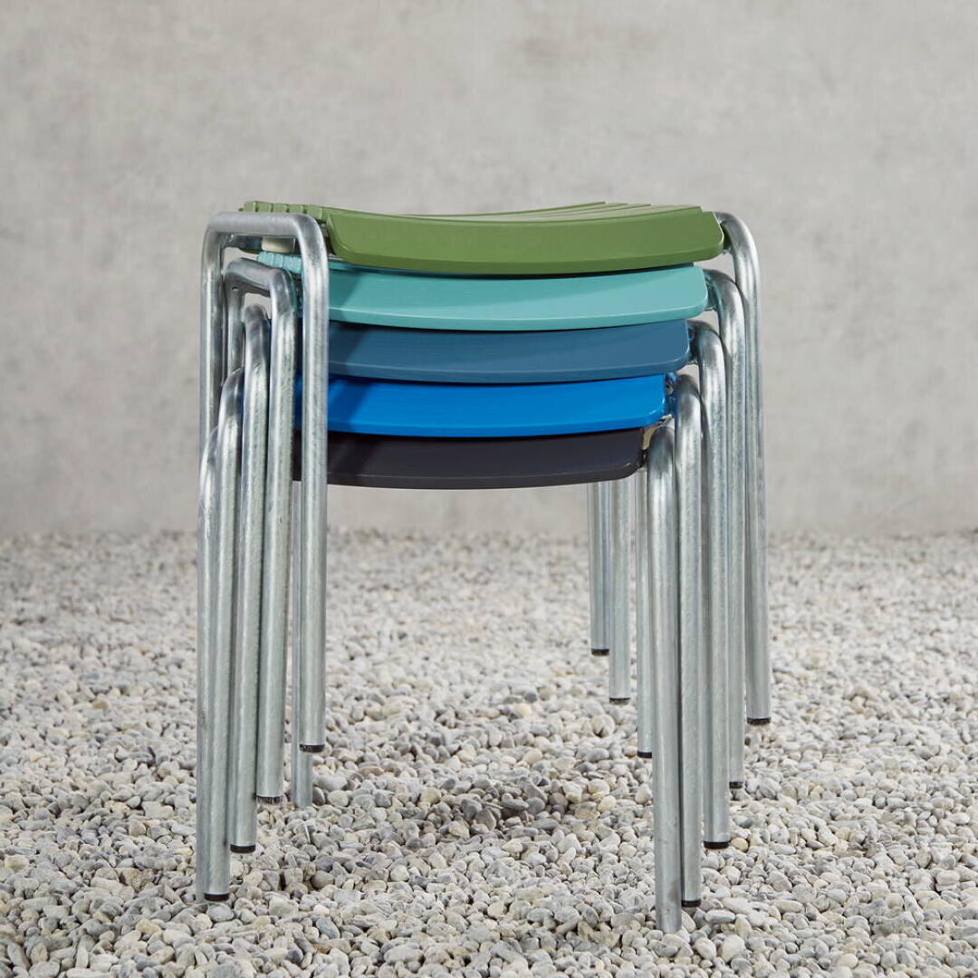 Vier stapelbare Hocker mit Metallbeinen und Kunststoffsitzen in den Farben Grün, Türkis, Türkis und Blau stehen auf einem Bett aus kleinen Kieselsteinen vor einem schlichten Betonhintergrund.
