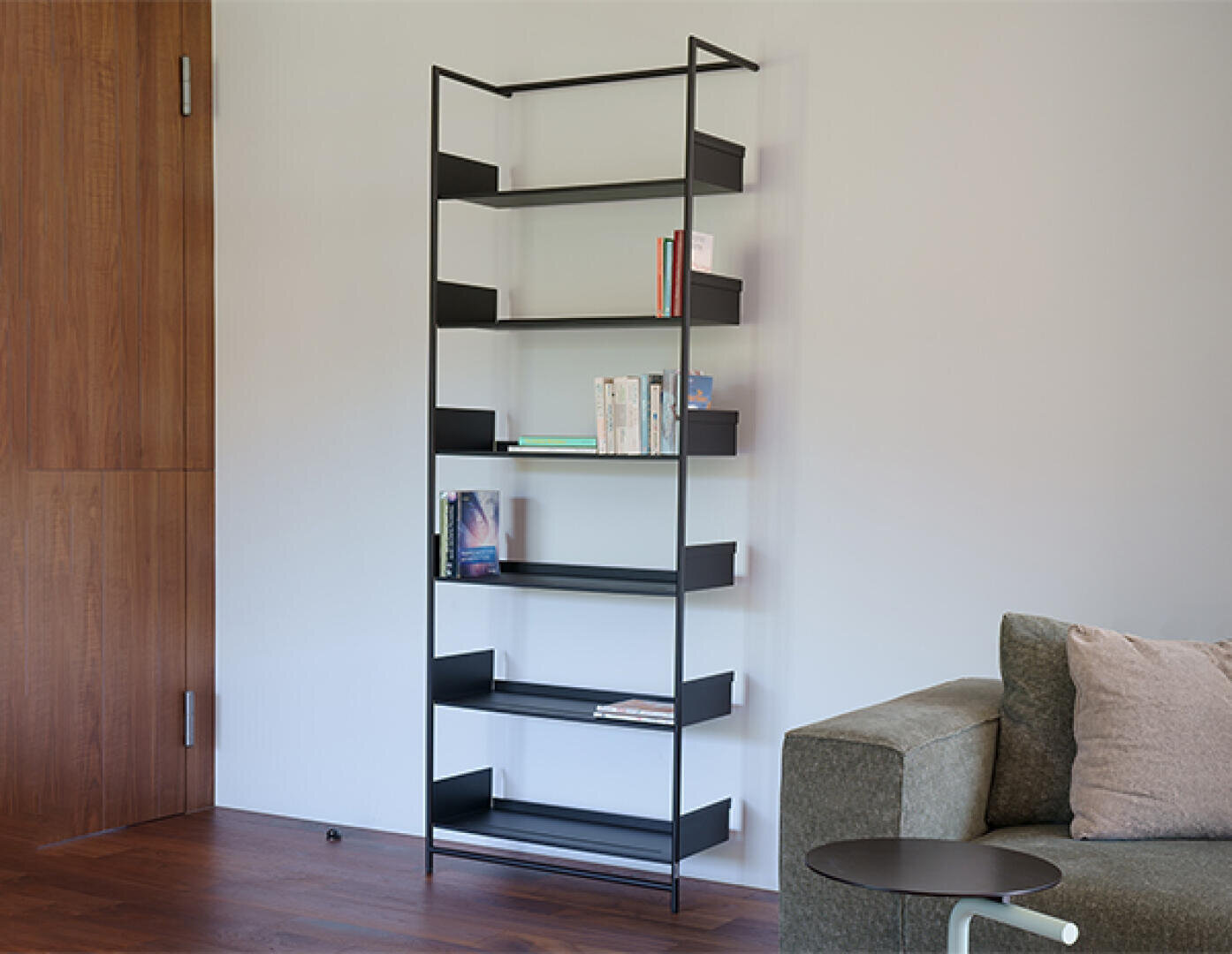 Ein hohes, schwarzes Metallregal mit einigen Büchern auf den Regalböden steht an einer hellgrauen Wand neben einem grünen Sofa und einem runden Beistelltisch in einem modernen Wohnzimmer mit Holzfußboden.