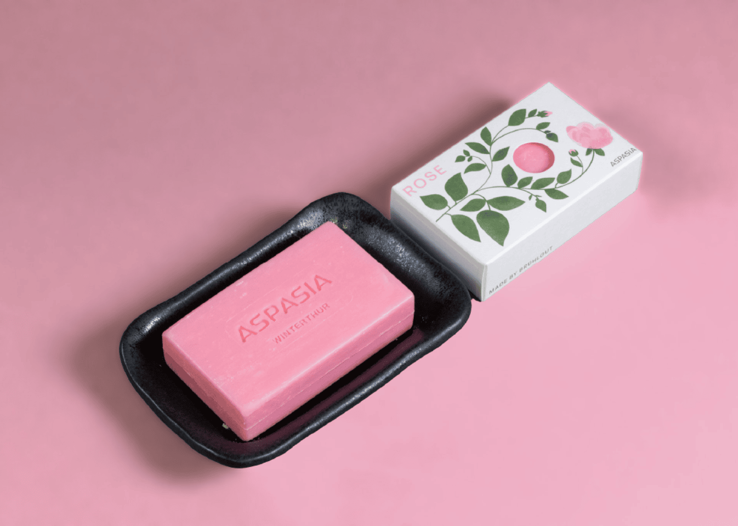 A pink bar of soap with the inscription ASPASIA lies on a black dish next to a white soap box decorated with green leaves and a pink rose, all against a pink background.