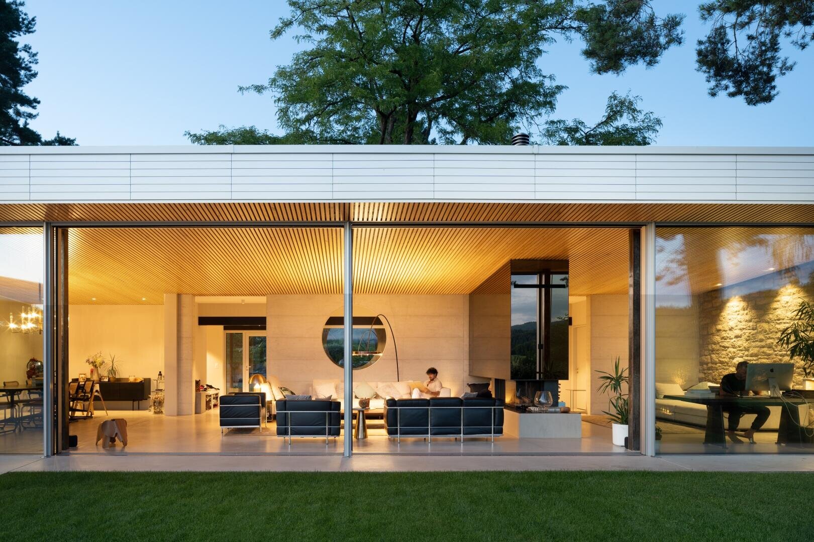 A modern house with large glass walls shows people relaxing inside on sofas and at a desk. The warm interior lighting contrasts with the evening sky outside, and a dog walks near the open living area.