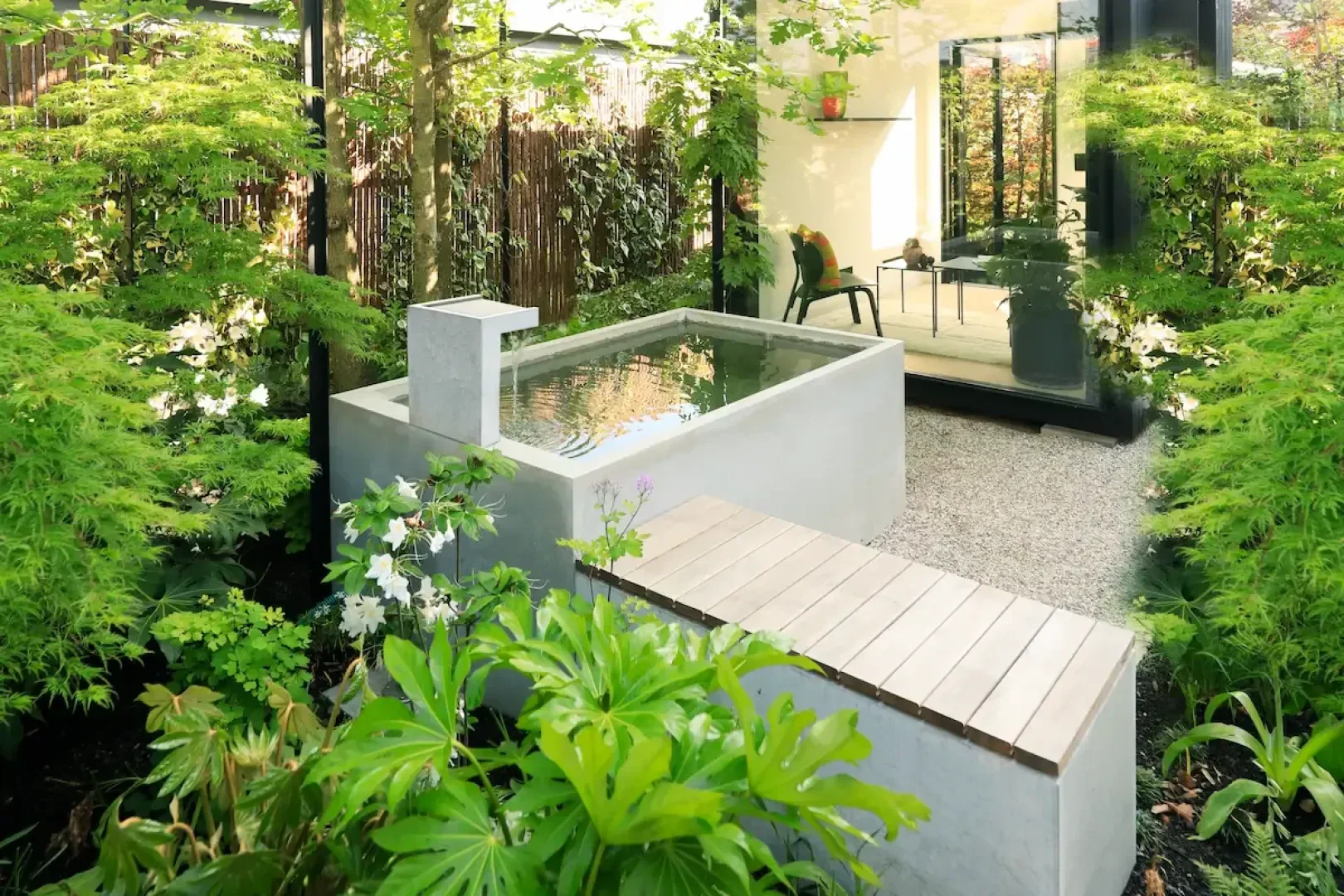 Ein moderner Garten im japanischen Stil mit einem kleinen rechteckigen, spiegelnden Pool, umgeben von üppigen Grünpflanzen und einer Holzbank. Eine Glastür führt in einen Innenraum, in dem ein Stuhl steht.
