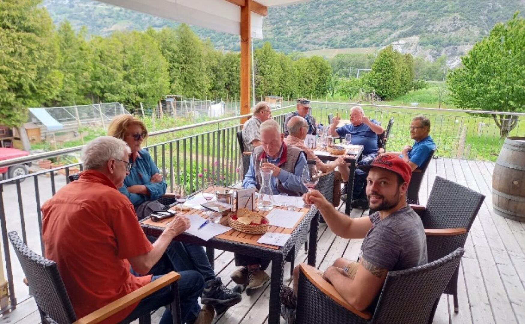 A group of people sit around a table on a covered outdoor patio, enjoying drinks and conversation. The background features lush green trees and hills, creating a relaxed and scenic atmosphere.