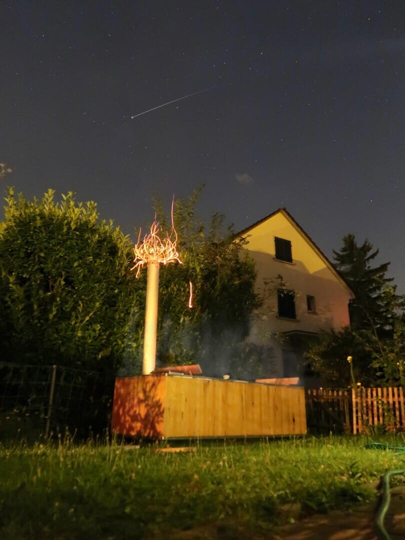 A wooden outdoor grill emits glowing sparks and smoke at night in a backyard. Behind the grill, a house and trees are visible under a starry sky with a shooting star.