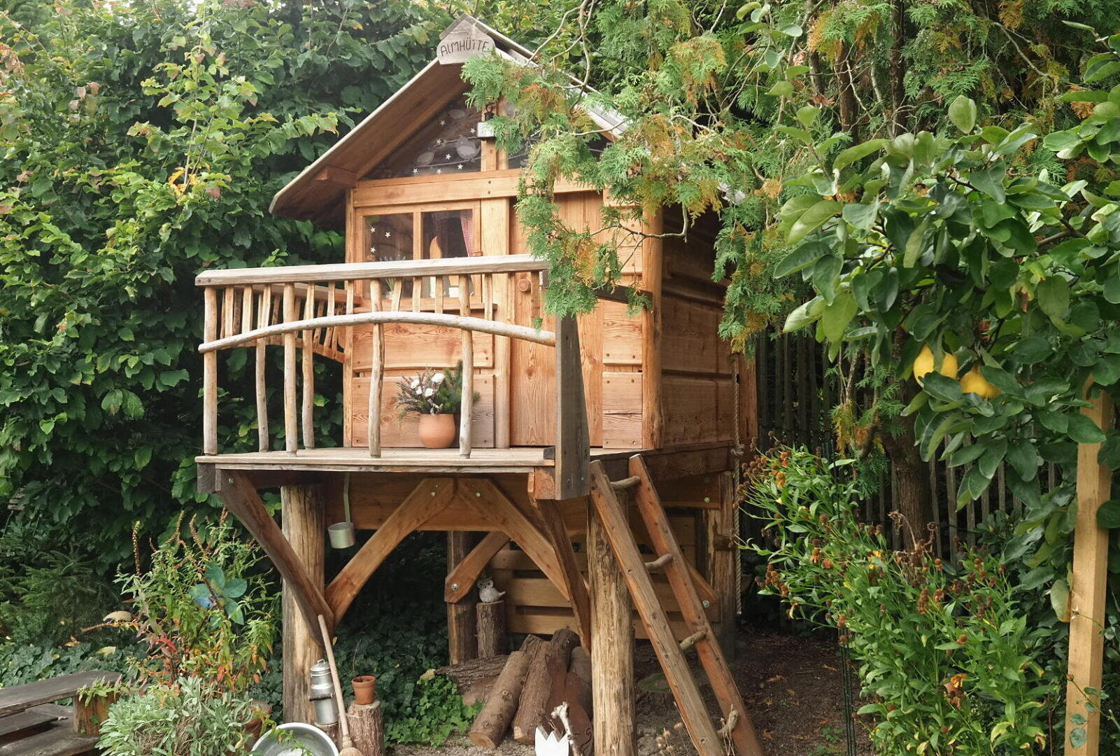Ein kleines hölzernes Baumhaus mit einem Balkon, einer Topfpflanze und einer Leiter steht zwischen grünen Bäumen und Büschen in einem Garten. Darunter sind Baumstämme und Gartengeräte zu sehen.