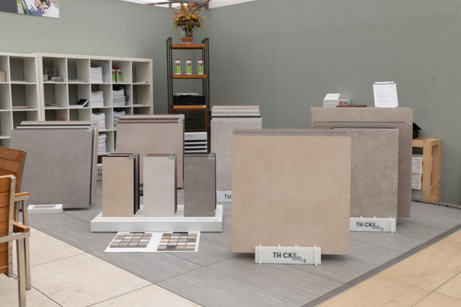 Große und kleine Keramikfliesenmuster in neutralen Farbtönen werden aufrecht auf Ständern in einem Ausstellungsraum mit Regalen, Stühlen und Präsentationsständern im Hintergrund ausgestellt.