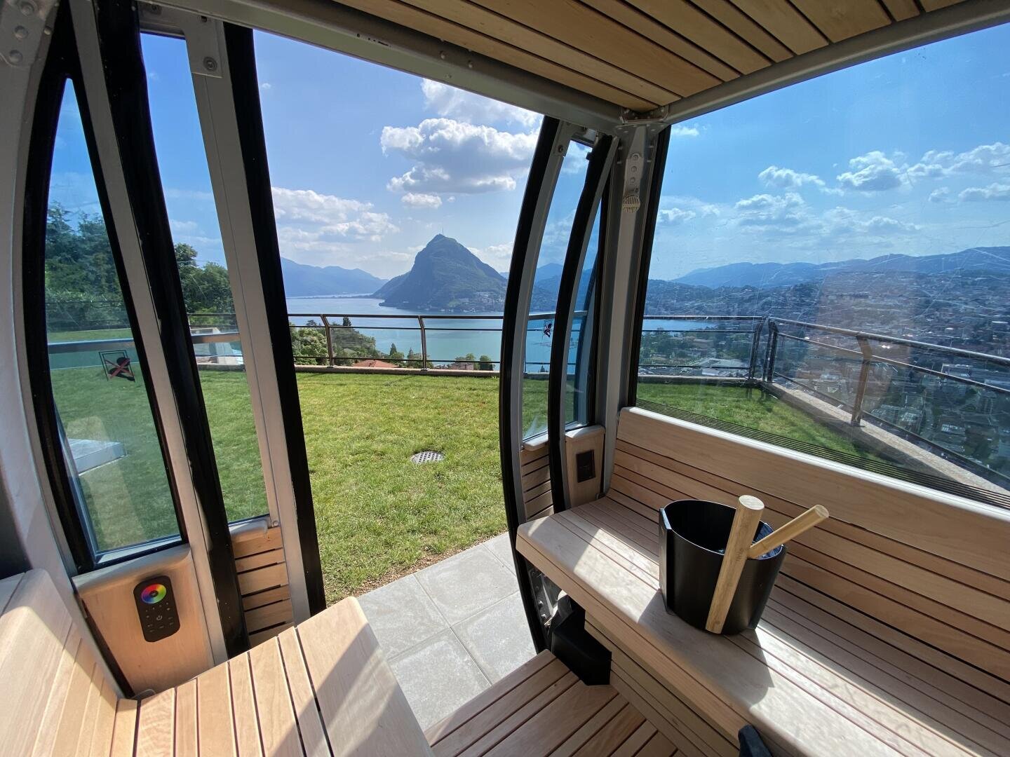Blick aus dem Inneren einer modernen Holzsauna mit großen Fenstern auf eine grüne Wiese, einen See und Berge bei teilweise bewölktem Himmel. Ein Saunakübel und eine Schöpfkelle stehen auf einer Bank.