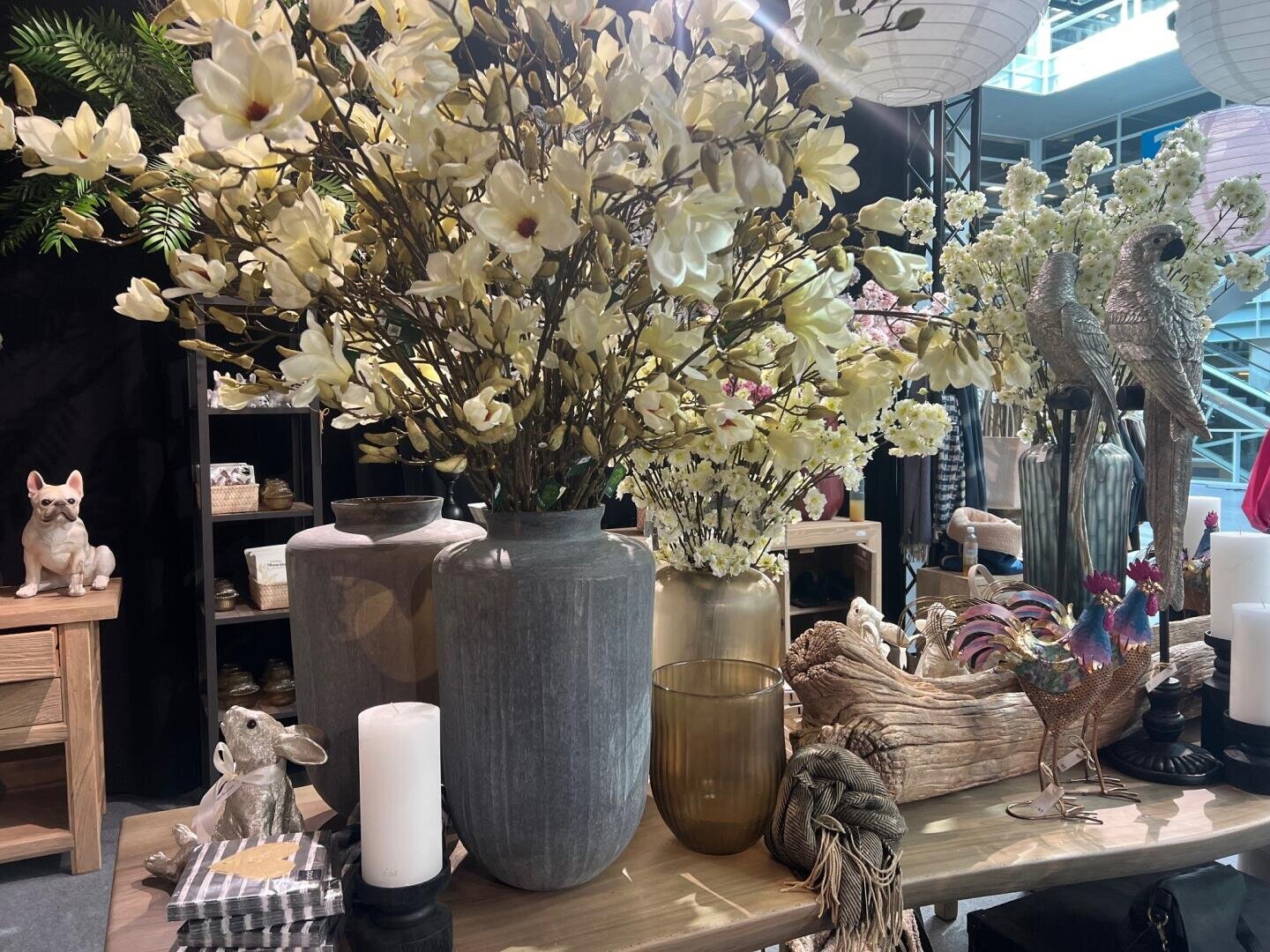 Ein Ausstellungstisch mit großen Vasen mit cremefarbenen Blumen, dekorativen Keramiktieren, Kerzen, einem hölzernen Hahn und anderen Dekorationsartikeln in einem gut beleuchteten Innenraum.