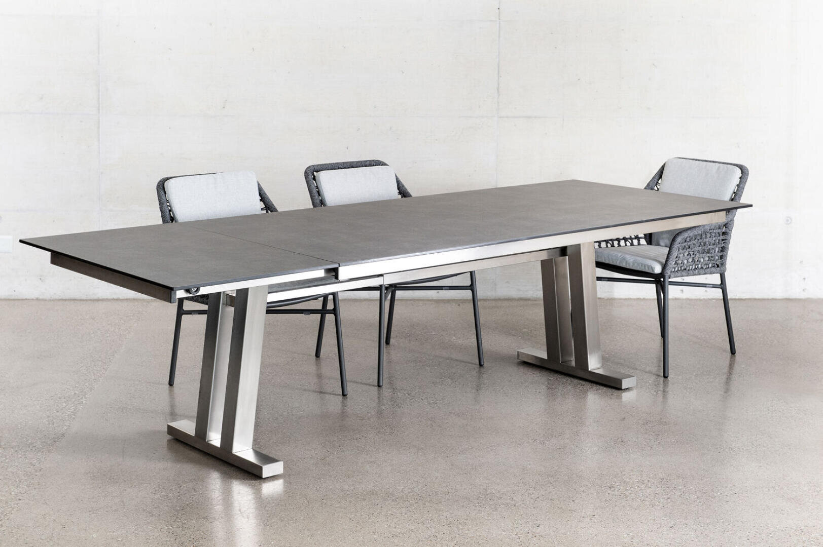 Ein moderner rechteckiger Esstisch mit dunkler Oberfläche und Metallbeinen wird von drei geflochtenen Stühlen mit hellen Polstern umgeben, die vor einer hellen Wand und einem polierten Betonboden stehen.