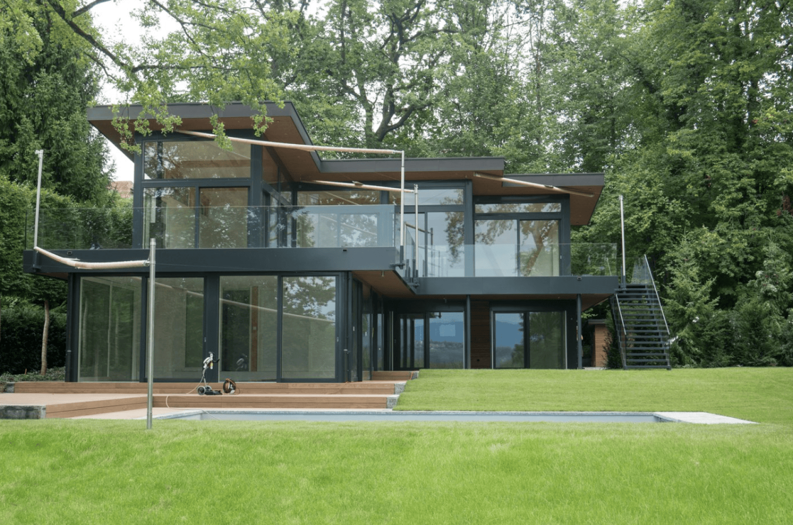Ein modernes zweistöckiges Haus mit großen Glasfenstern und Flachdächern, umgeben von üppigen grünen Bäumen und Gras, mit einer großen Rasenfläche und einem Swimmingpool im Vordergrund.