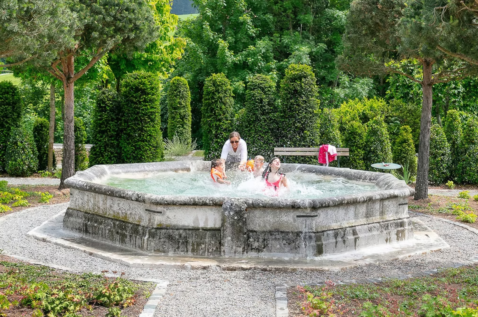 Four children and an adult splash and play in a large, octagonal stone fountain surrounded by greenery and trees in a park on a bright, sunny day.