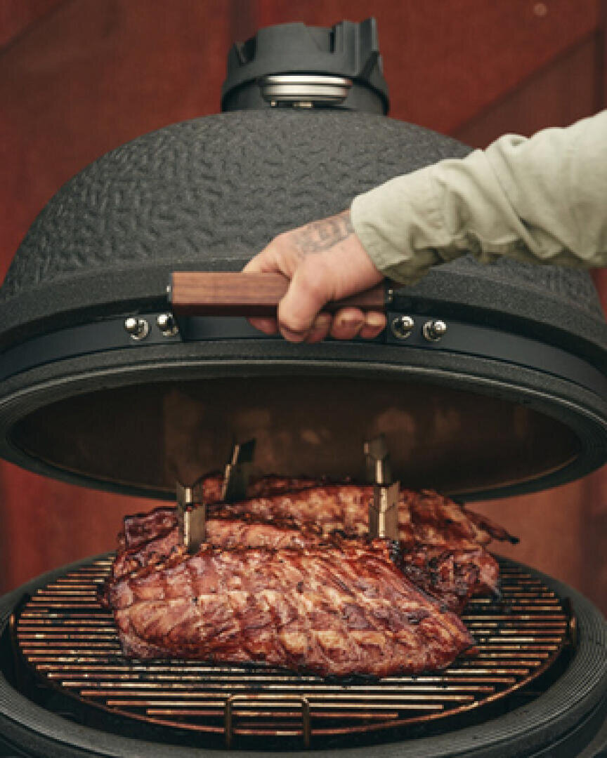 A person lifts the lid of a round, black grill and reveals the ribs being grilled on the grid. The grill has a textured surface and a wooden handle.