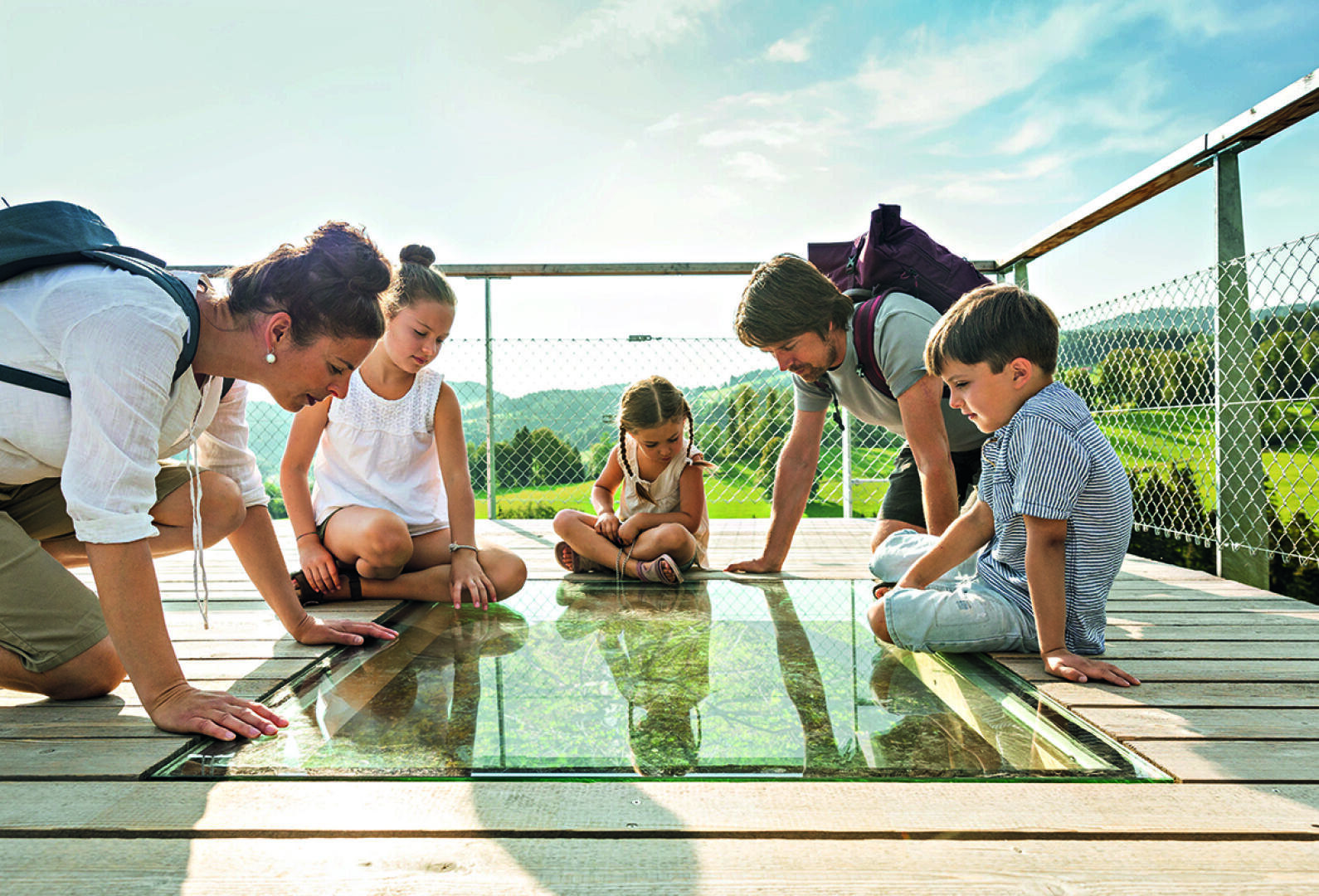 A family of five kneels on a wooden platform and looks through a glass floor at the landscape below, surrounded by a wire fence and green hills in the background.